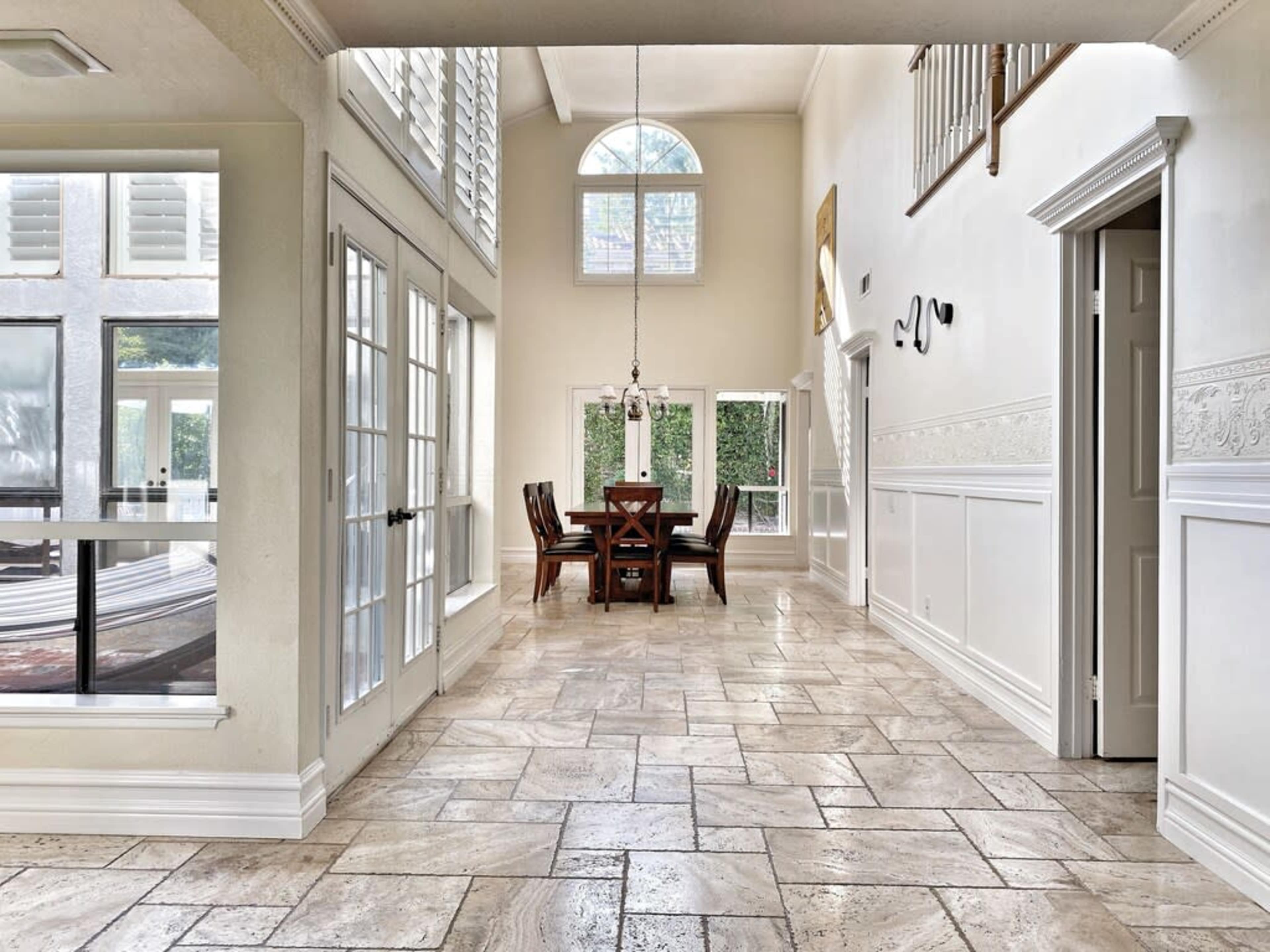 A spacious, brightly lit room with a large dining table surrounded by chairs, large windows, and tiled flooring.