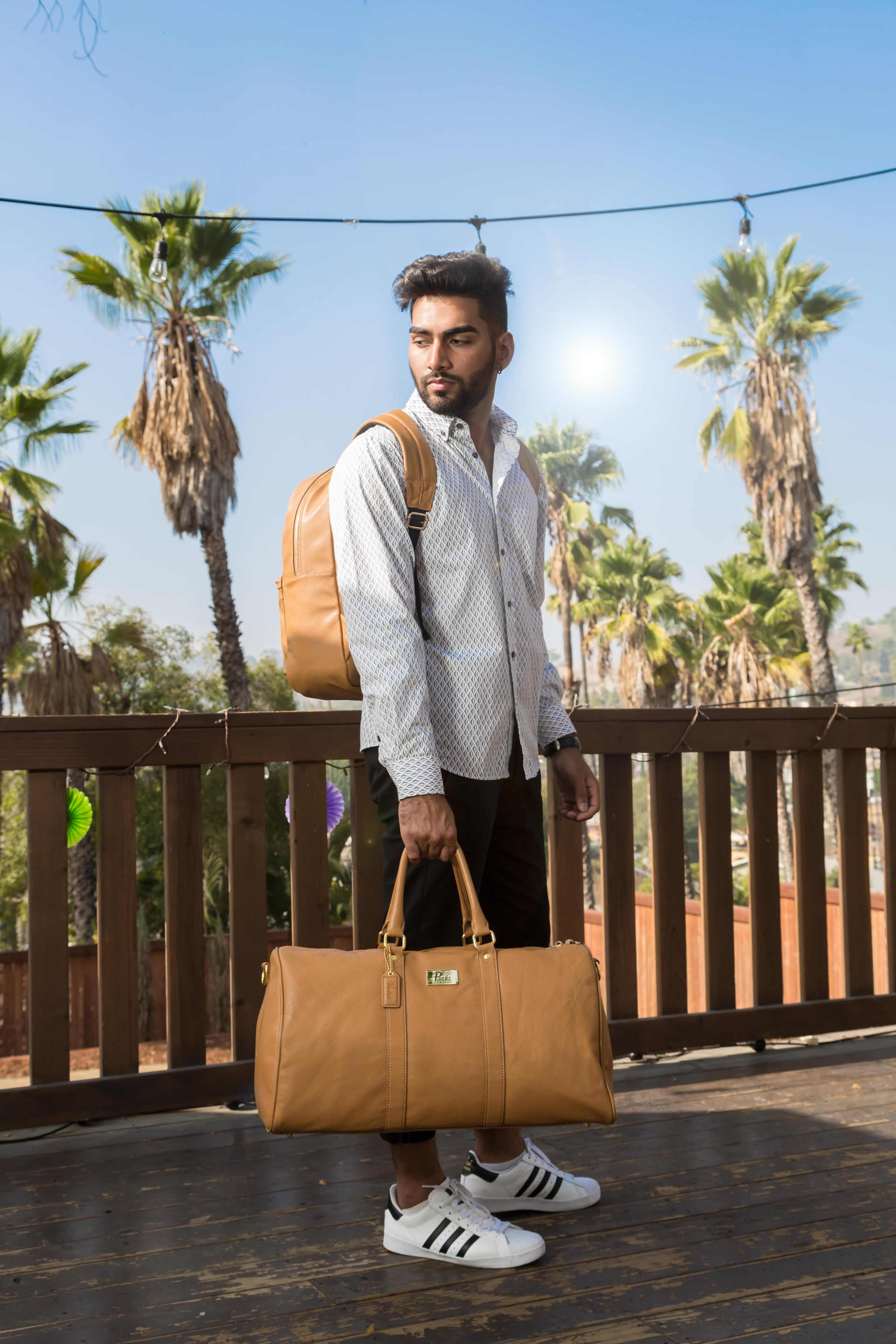 A man stands on a wooden deck with palm trees in the background, wearing a patterned shirt and carrying a backpack and a duffel bag.
