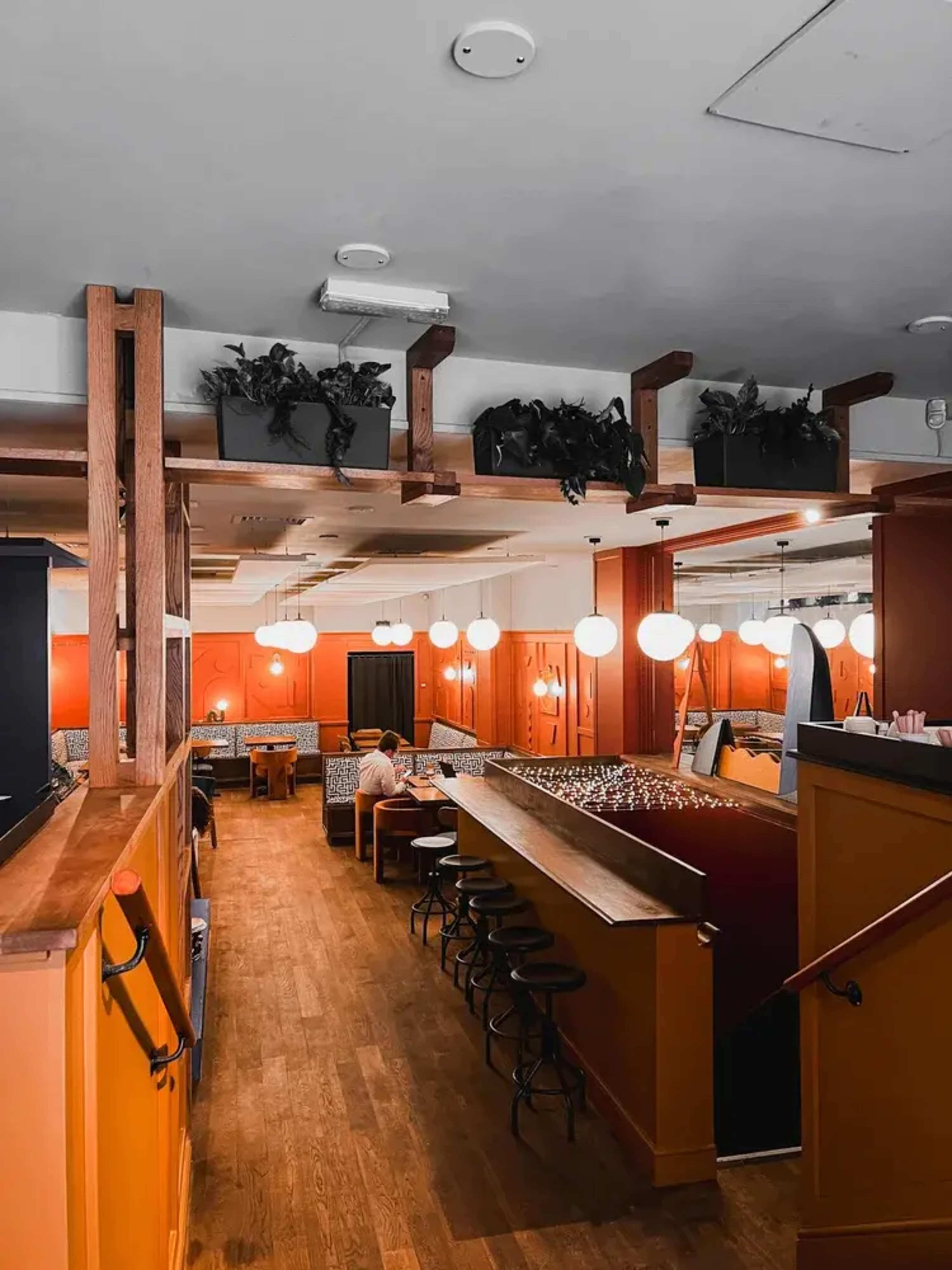 The image shows the interior of a restaurant featuring wooden flooring, round pendant lights, and a bar area with stools along one side.