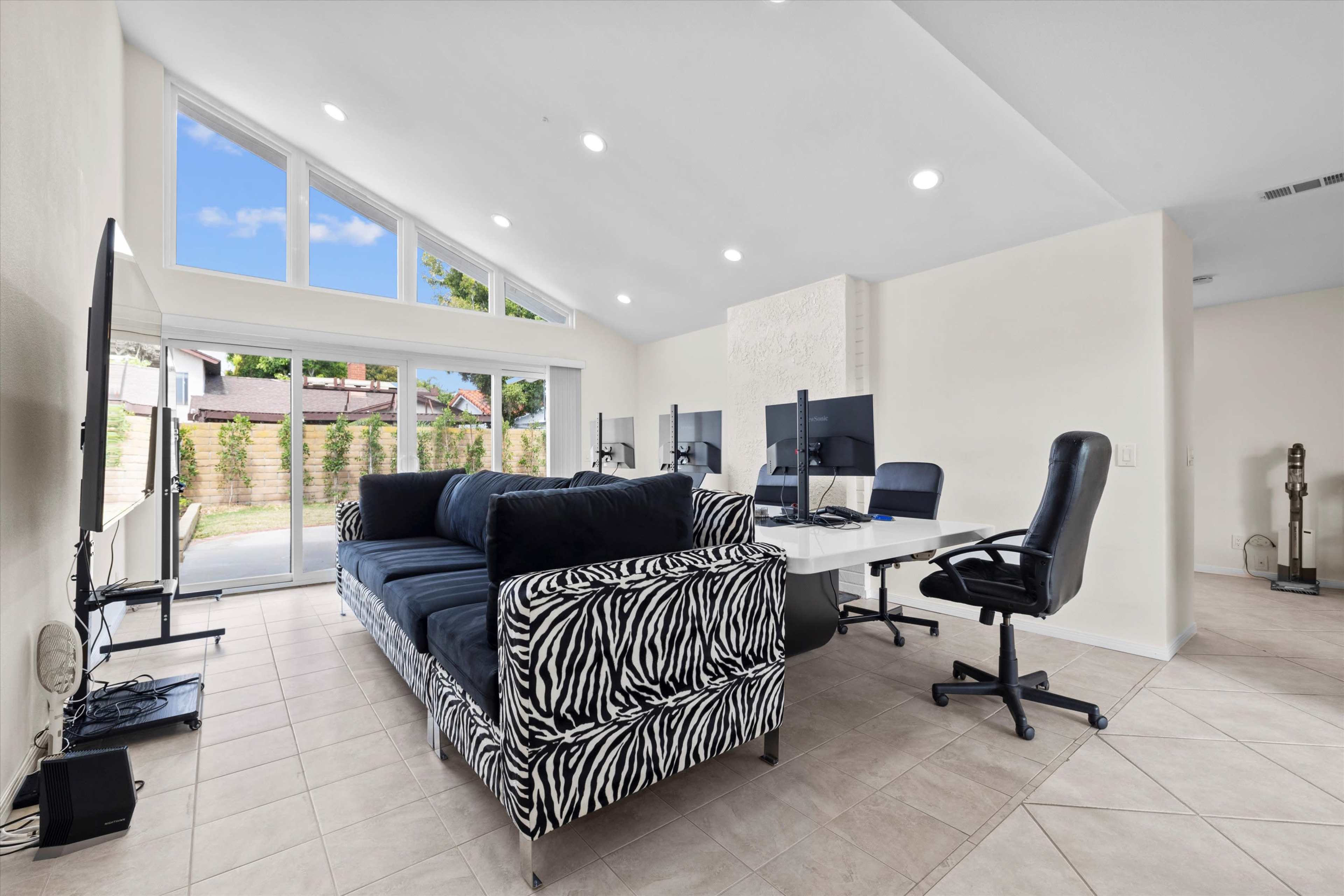 The image shows a spacious home office with a large zebra-patterned couch, multiple computer desks, and large windows allowing natural light.