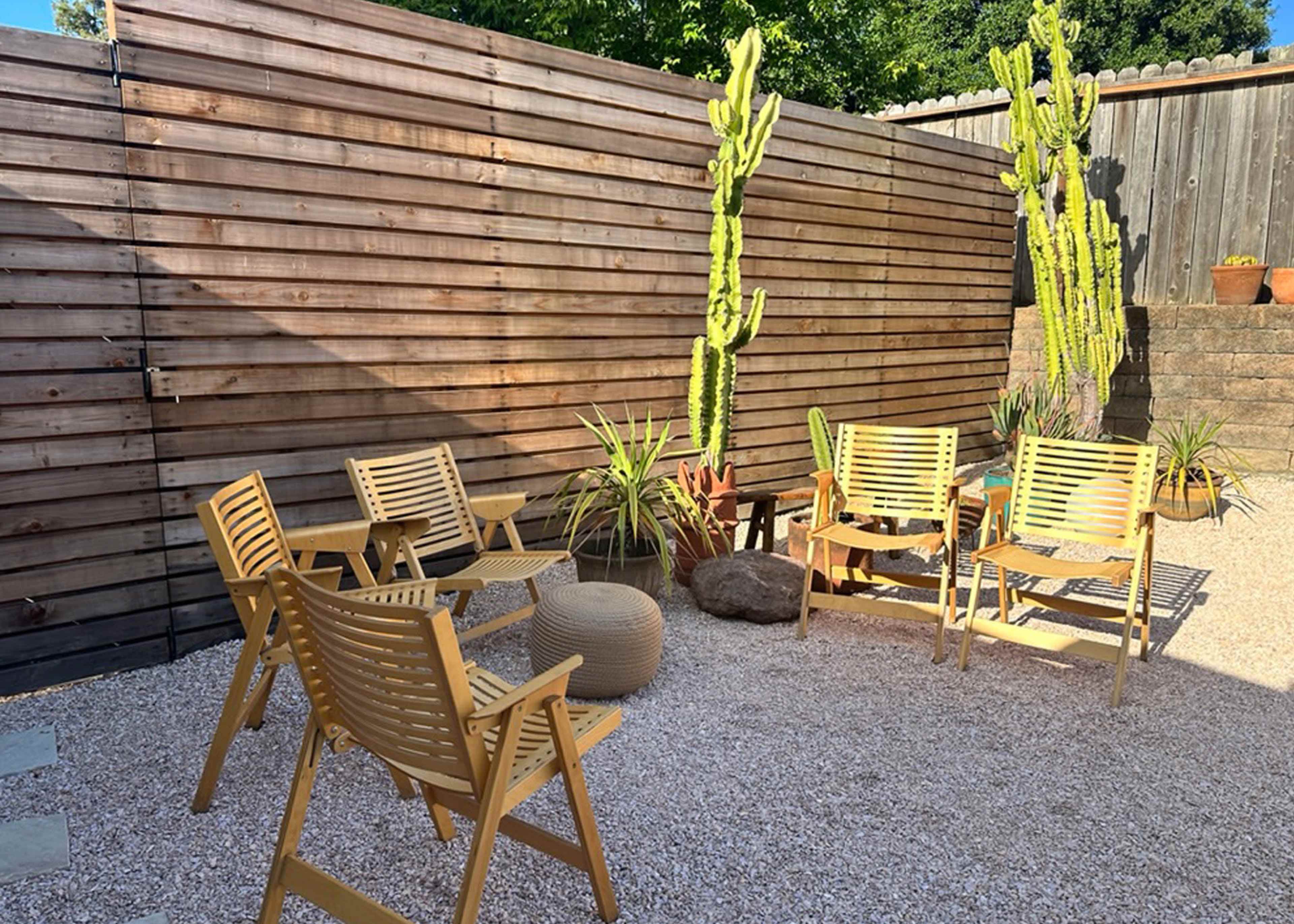 A small outdoor seating area with five wooden chairs arranged around a stone table, surrounded by gravel and cacti against a wooden fence.