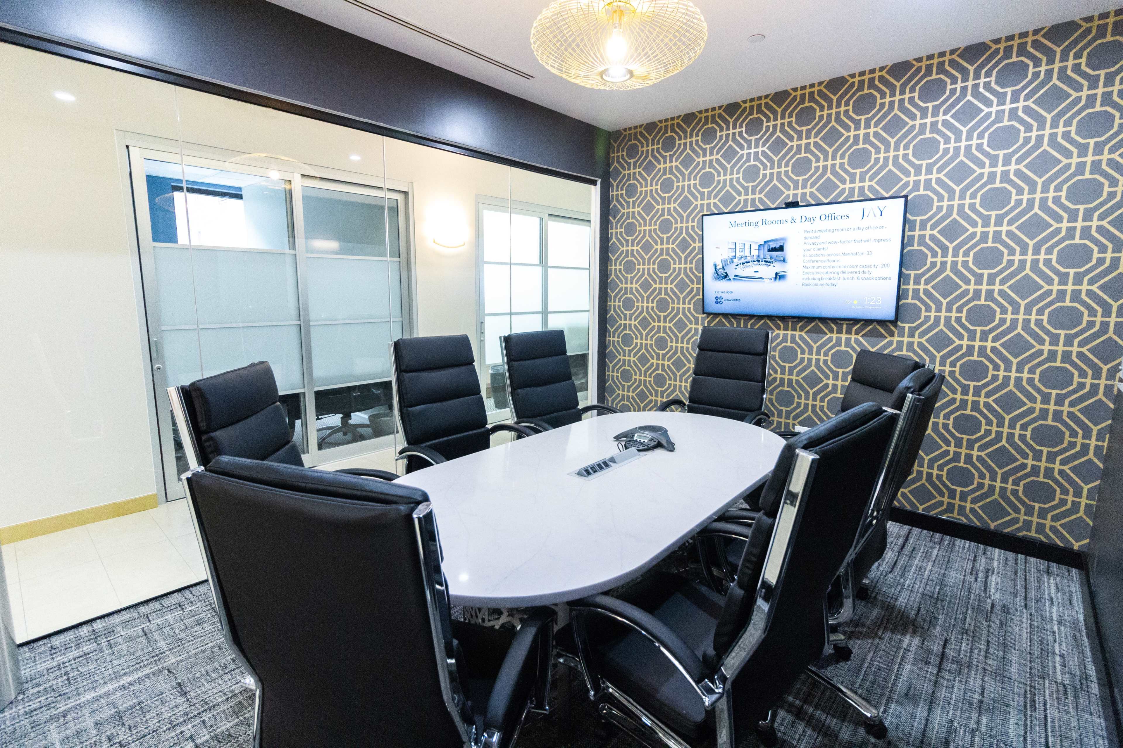 A modern conference room features a large oval table surrounded by black leather chairs, with a wall-mounted screen displaying information and a patterned wallpaper.