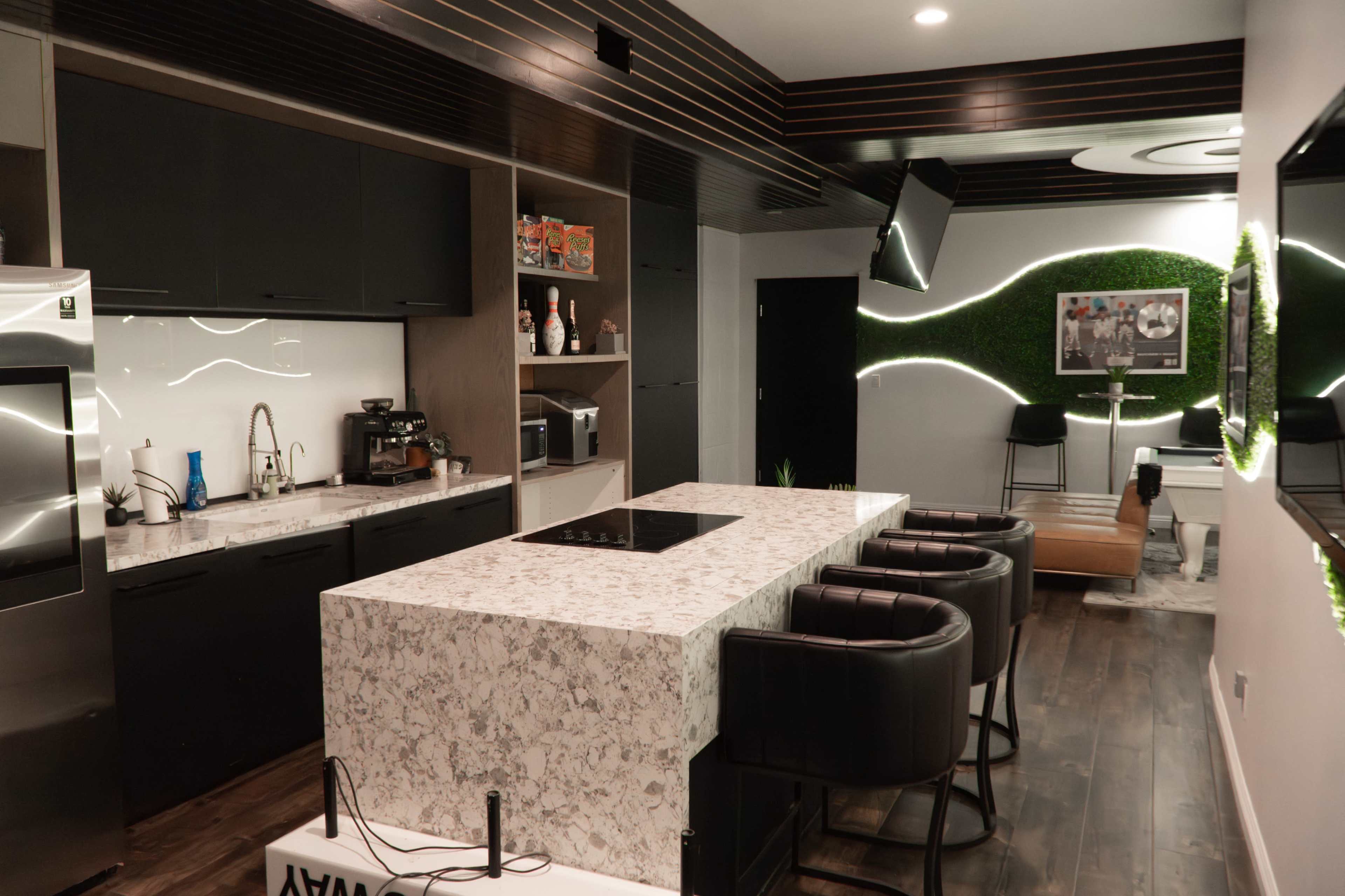 The image shows a modern kitchen with a large island, black cabinets, and a counter with bar stools, featuring various appliances and decorative elements.