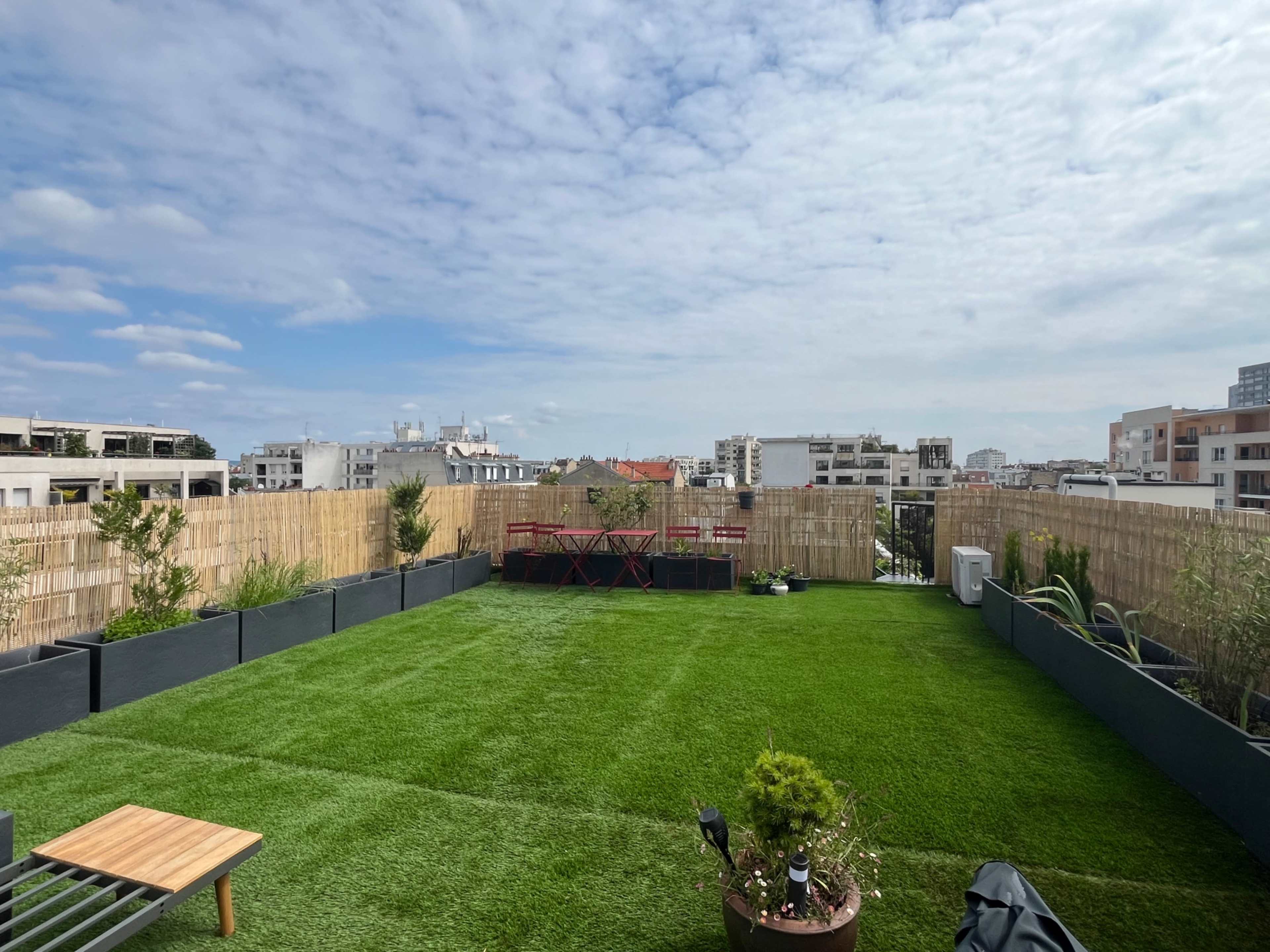 The image shows a spacious rooftop terrace featuring artificial grass, planters, and a seating area overlooking a city skyline.