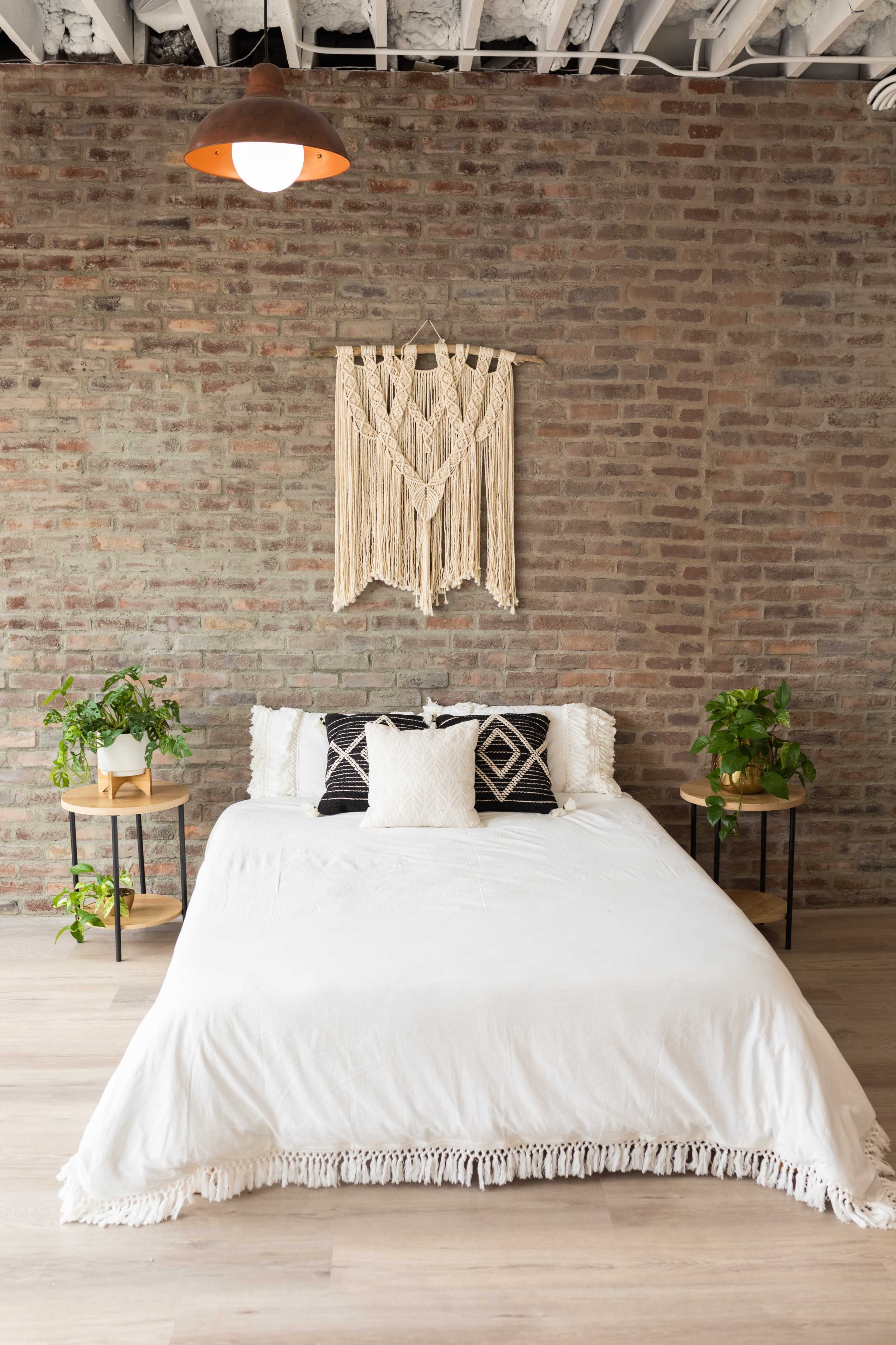A simple bedroom with a bed covered in a white blanket, two plant stands on either side, and a macramé wall hanging above the bed in front of a brick wall.