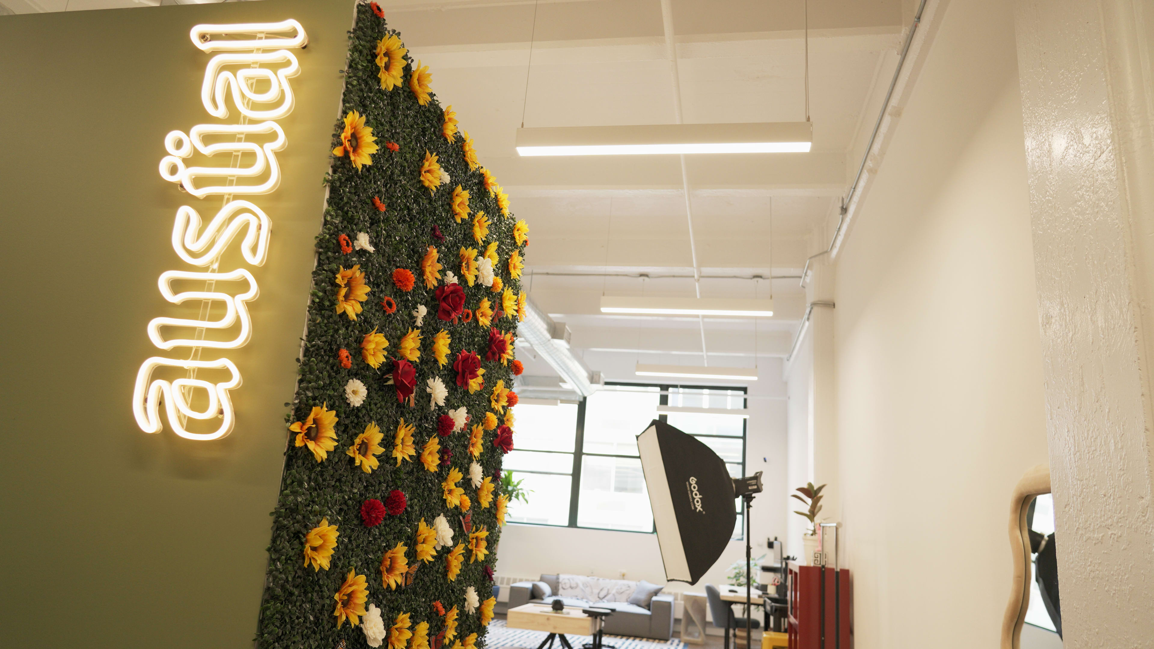 A wall covered in artificial greenery and colorful flowers features the word "austial" illuminated in neon lights, while a photography lighting setup is visible in a modern workspace.