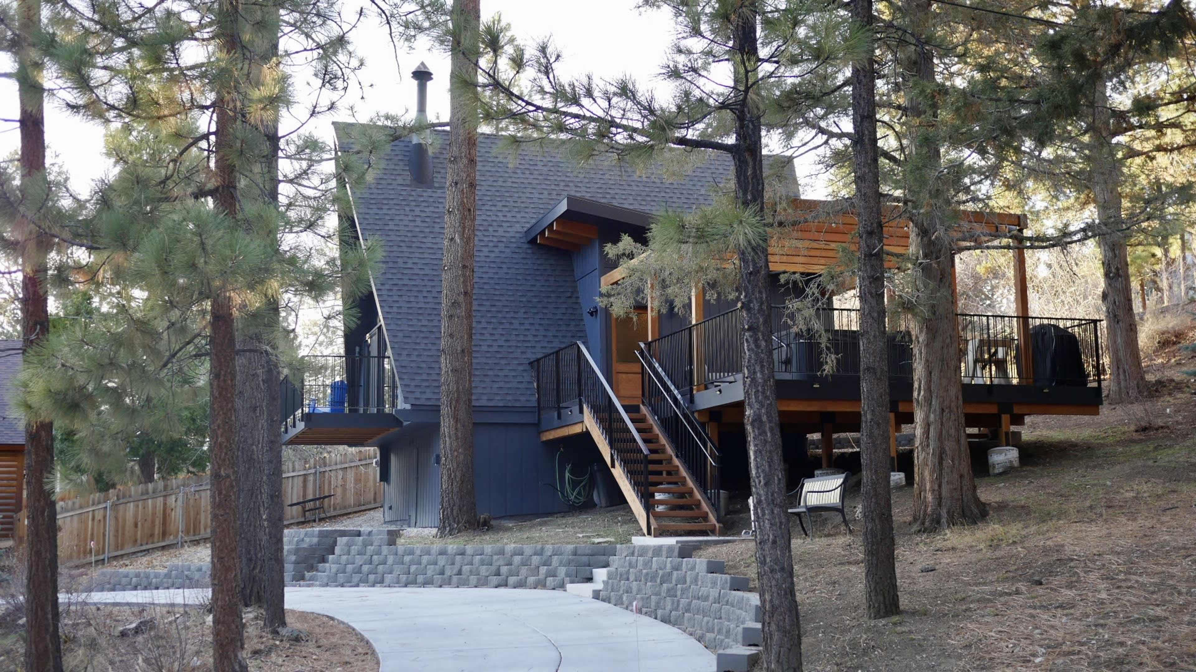 The image shows a two-story house with a deck, surrounded by tall pine trees, along a winding pathway.