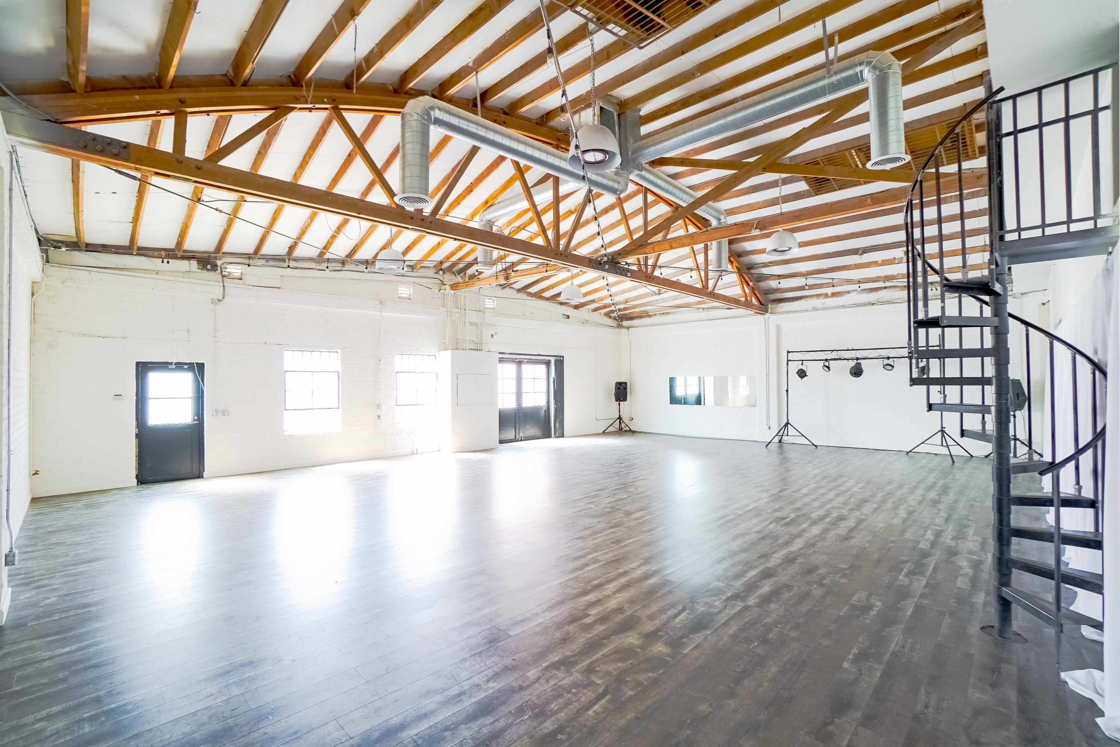 A spacious, empty room with wooden beams, large windows, and a spiral staircase in one corner.