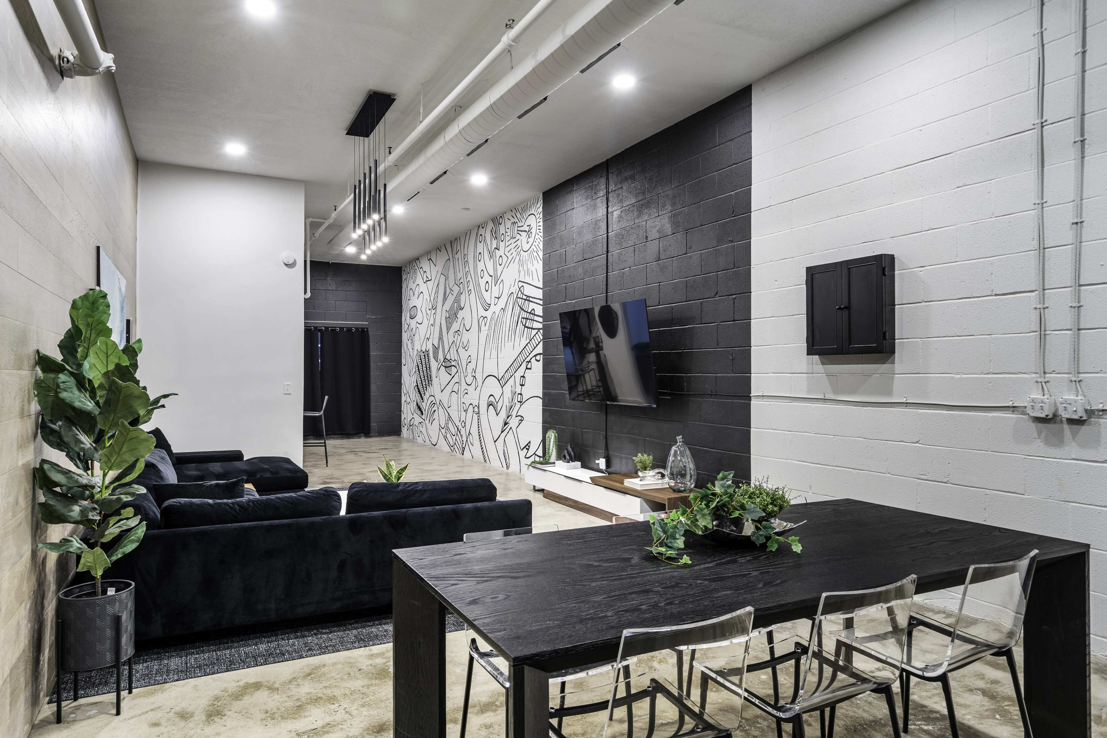 The image shows a modern interior space featuring a dining table with clear chairs, a black sofa, and a wall adorned with a mural.
