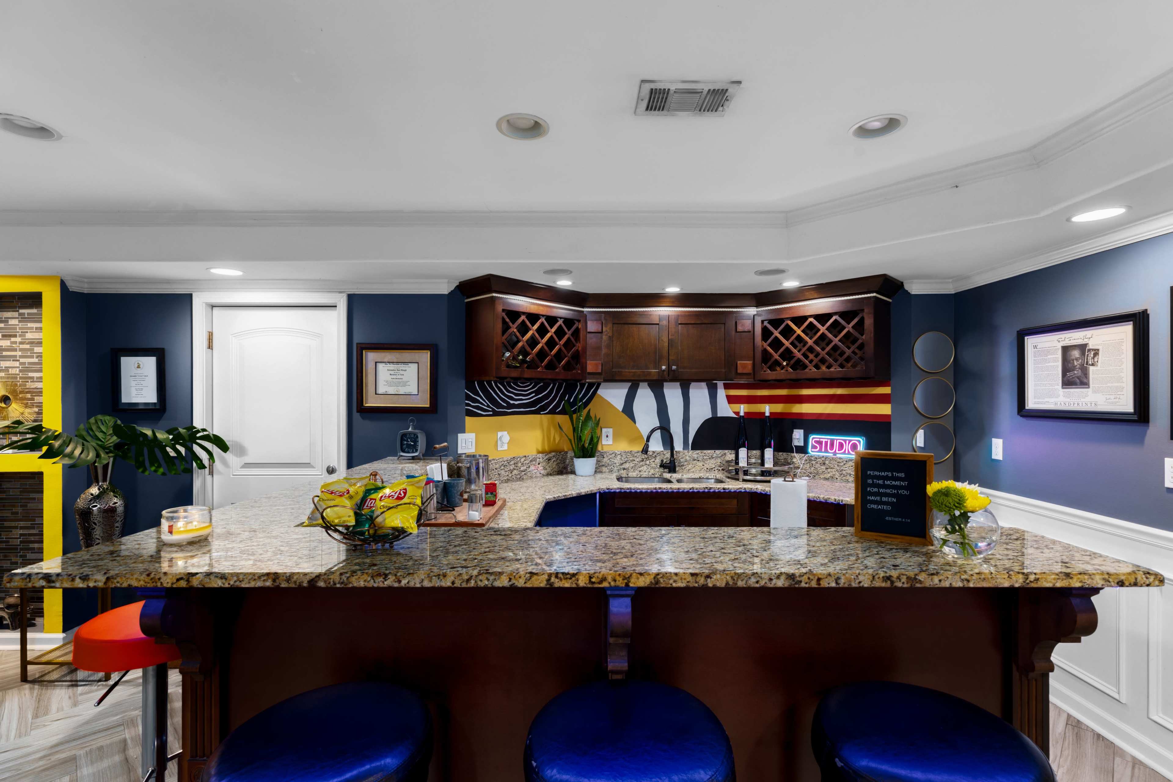 The image shows a modern kitchen bar with granite countertops, dark wooden cabinetry, and a variety of colorful decorations on the walls.