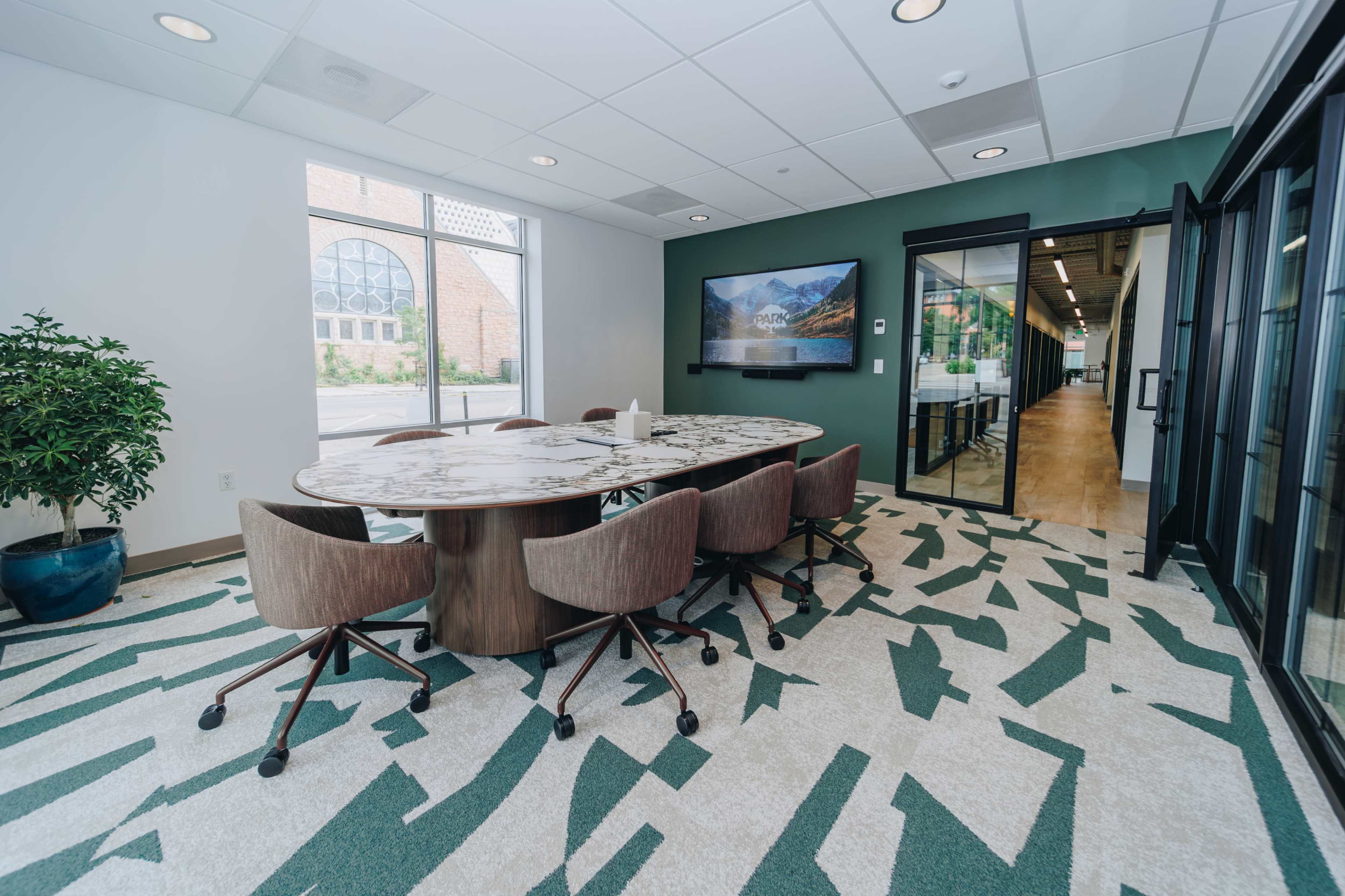 A modern conference room features a large, round table surrounded by upholstered chairs, with green and beige patterned flooring and a large window letting in natural light.