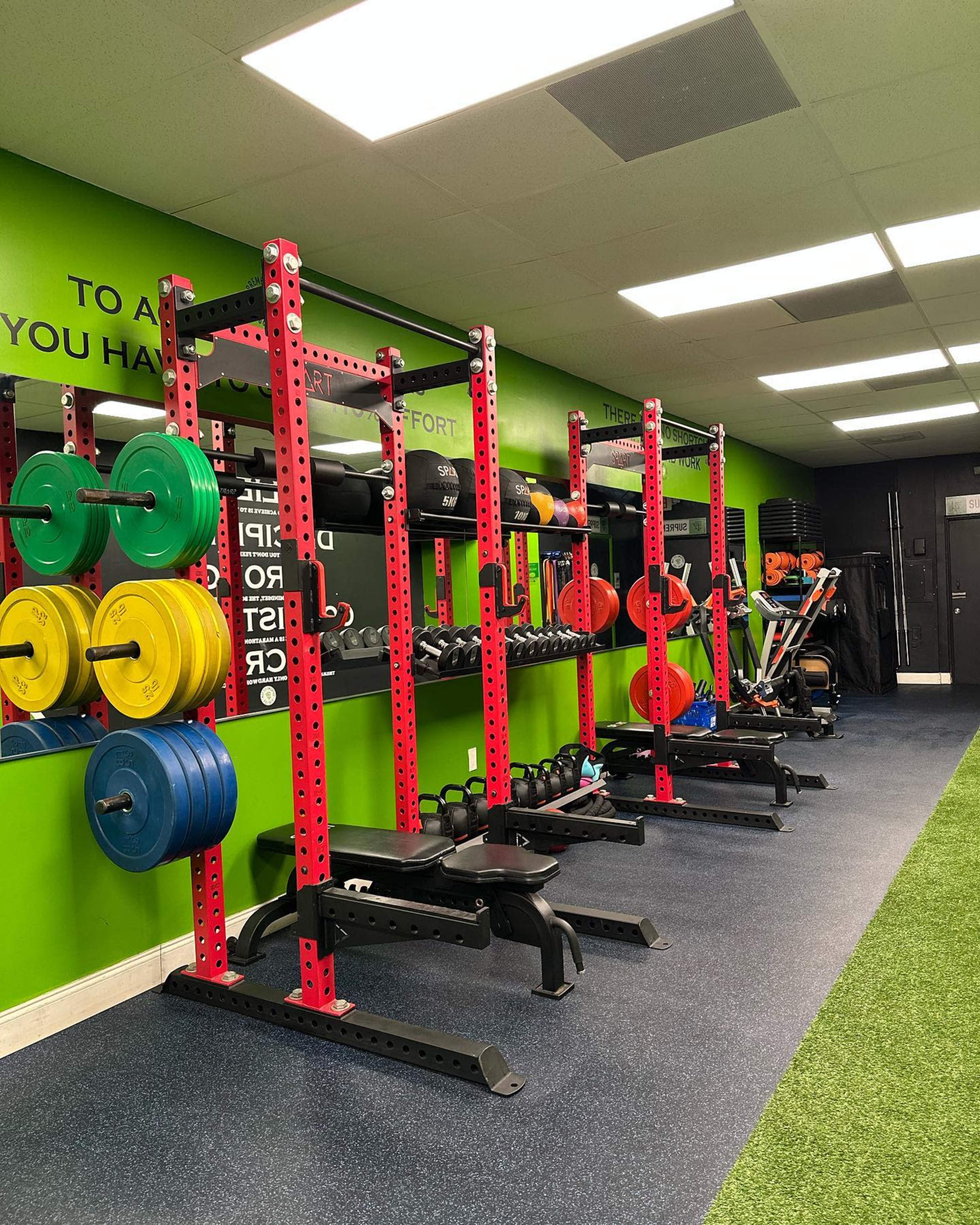 A gym interior with red weight racks, colorful plates, dumbbells, and fitness equipment arranged against a bright green wall.