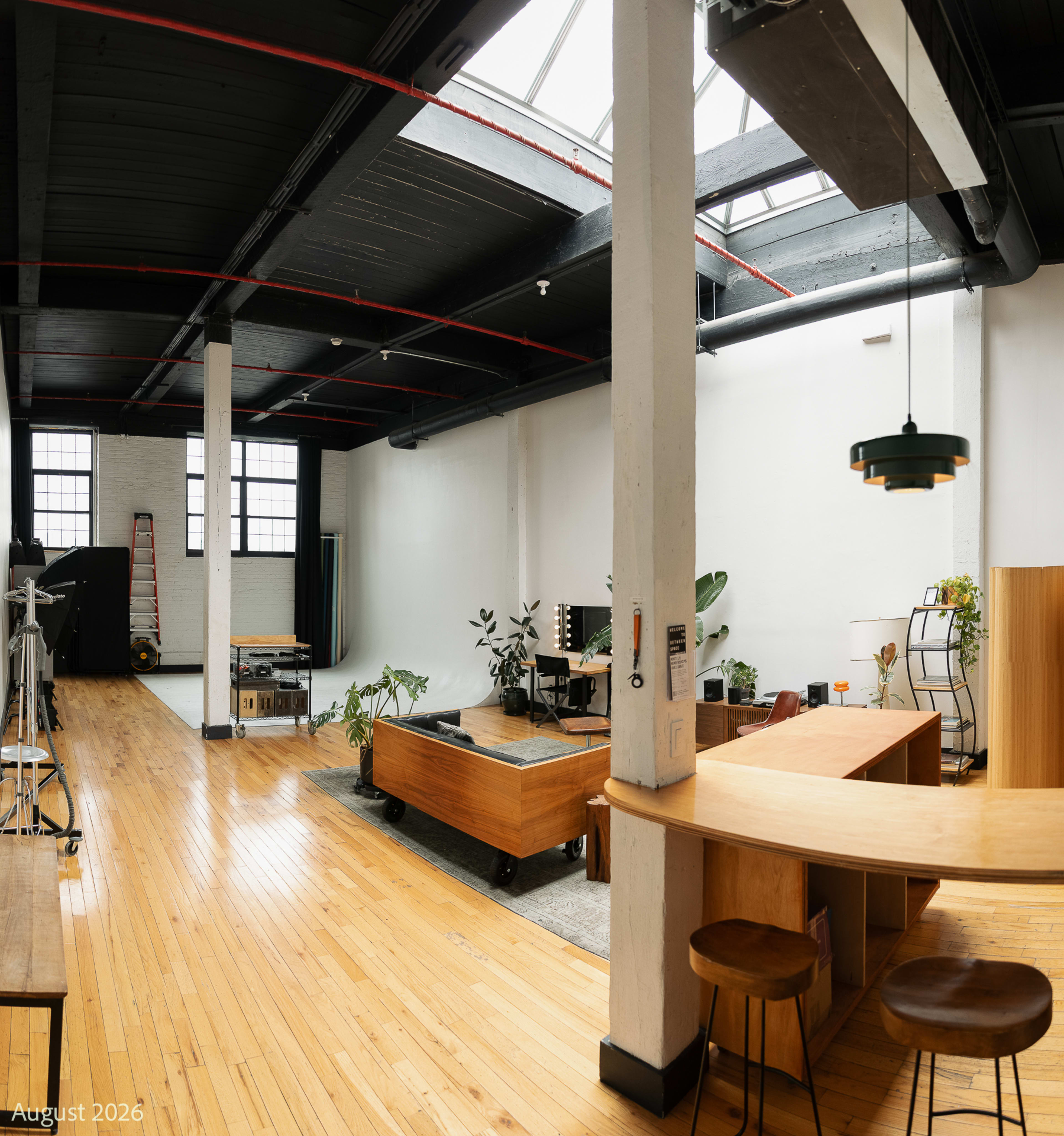 The image shows a spacious, well-lit studio with wooden flooring, large windows, and various furniture pieces including a bar counter, seating, and plants.
