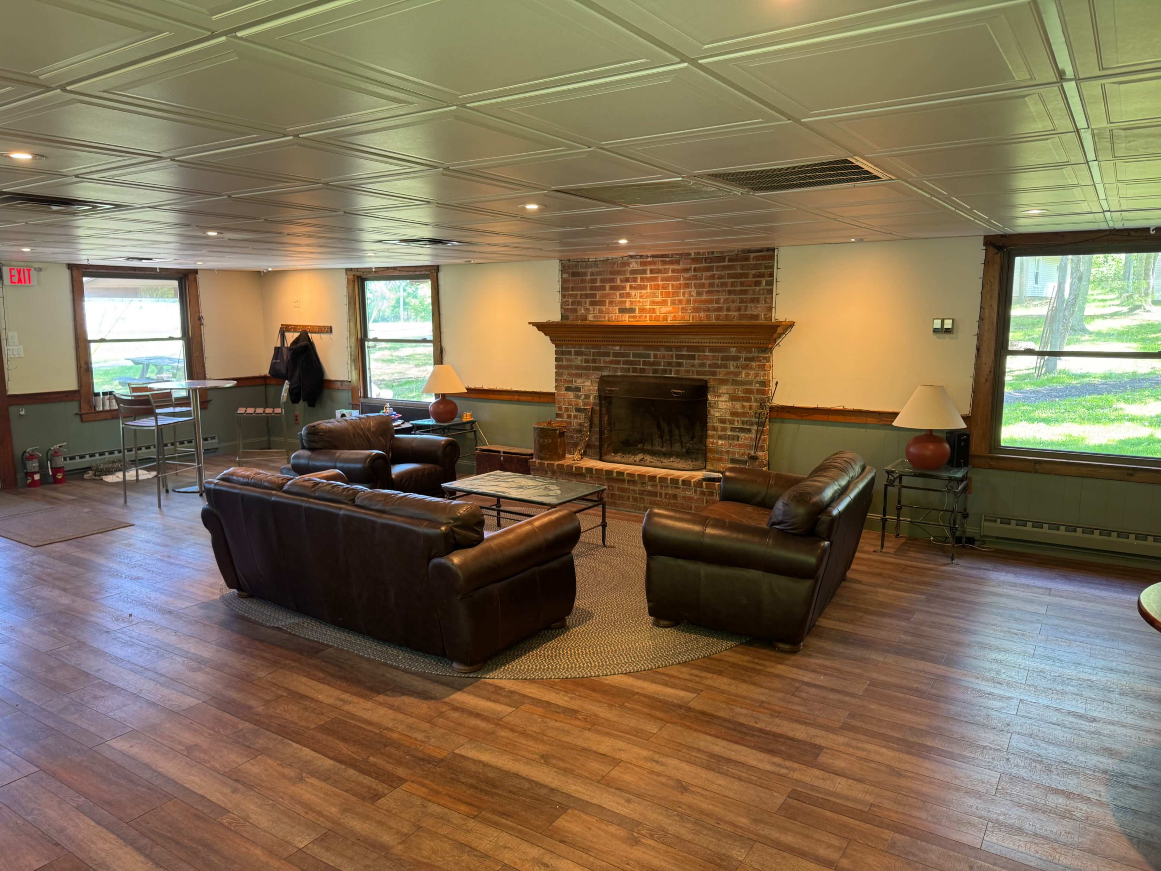 The image shows a spacious indoor lounge area featuring two leather sofas arranged around a brick fireplace, with large windows overlooking a green outdoor space.