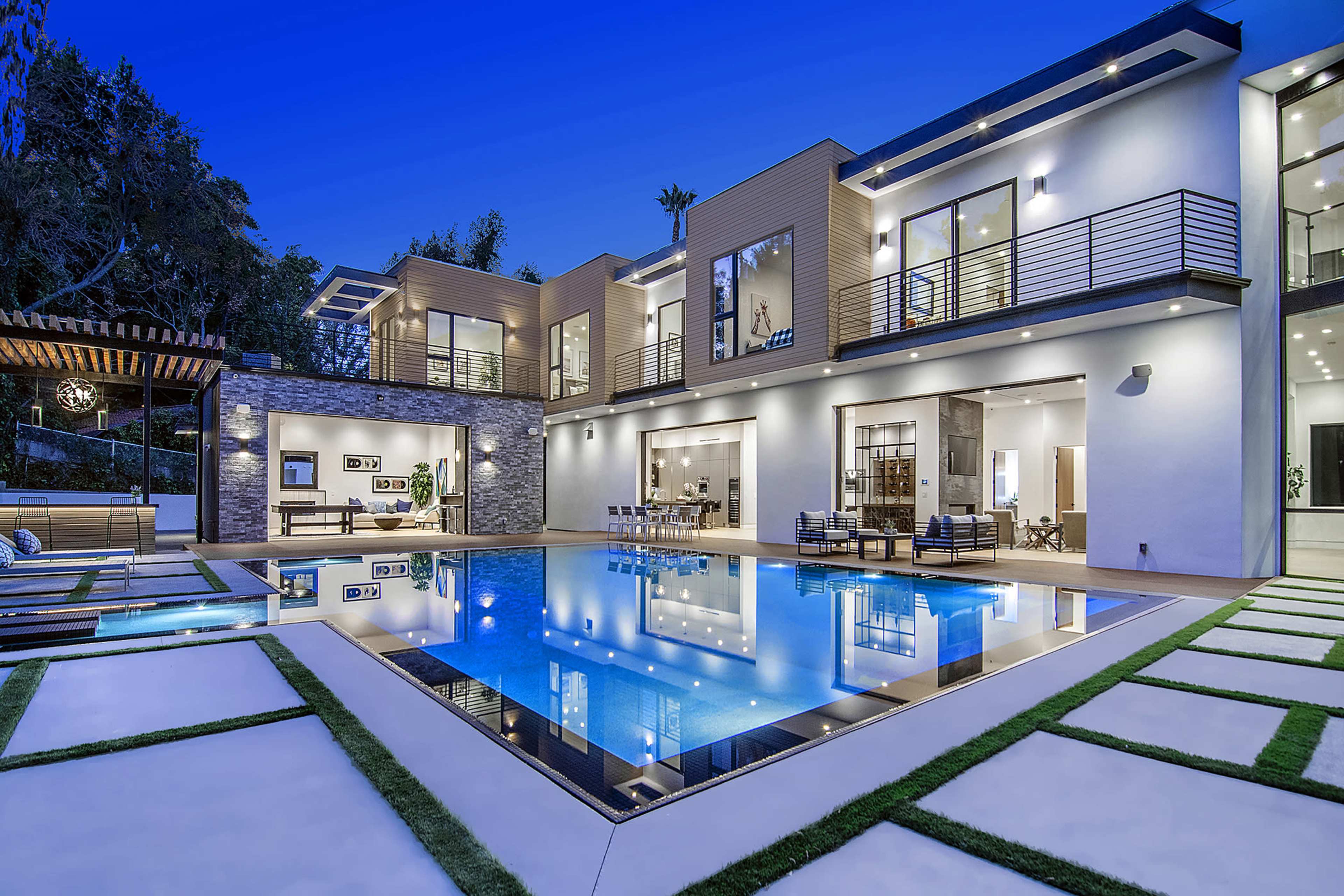 A modern house with large windows and a spacious pool area is illuminated at dusk, reflecting the surrounding landscape and architecture.