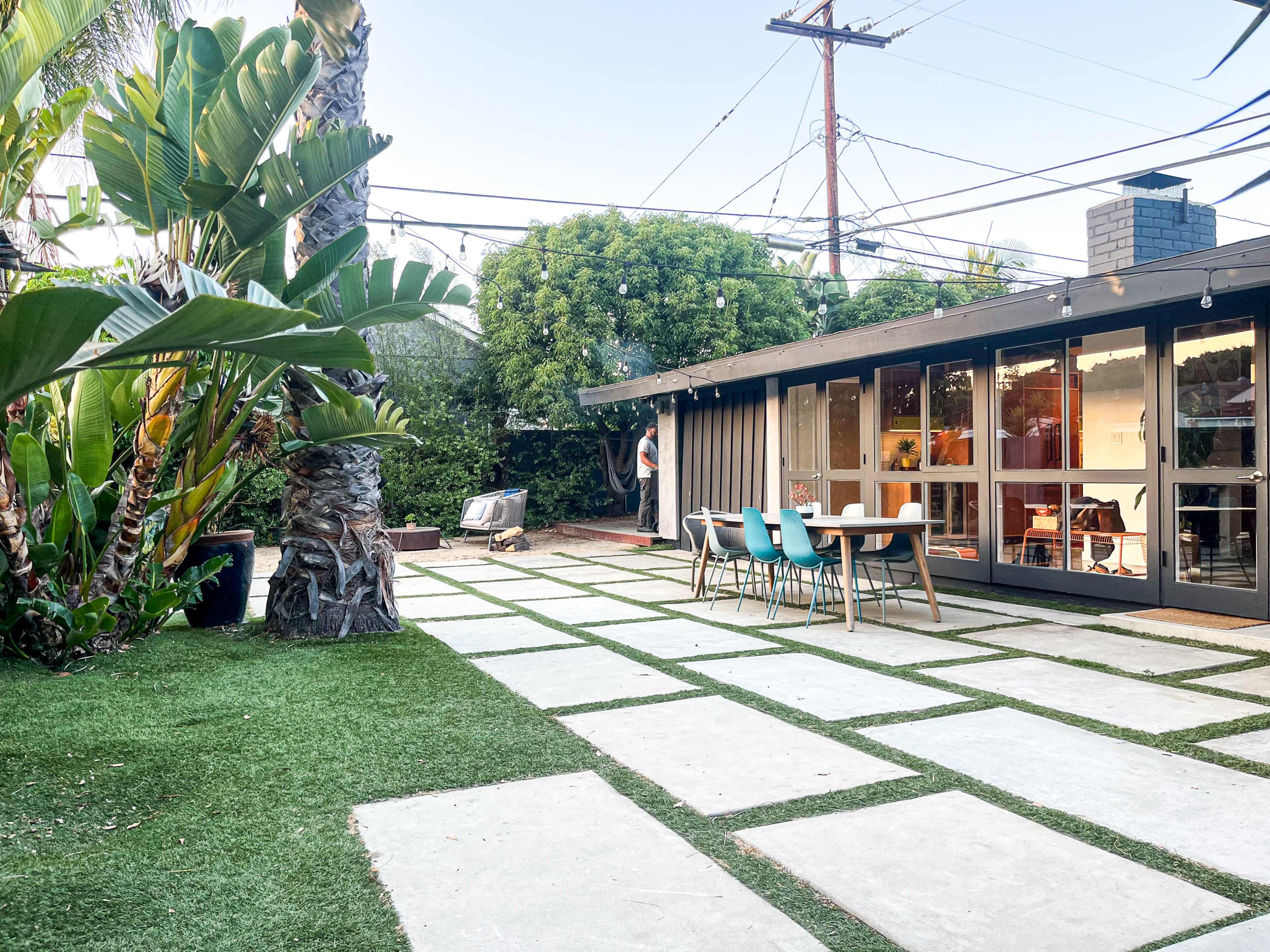 A modern backyard features a patio with a dining table, surrounded by greenery and decorative plants, adjacent to a small building with large windows.