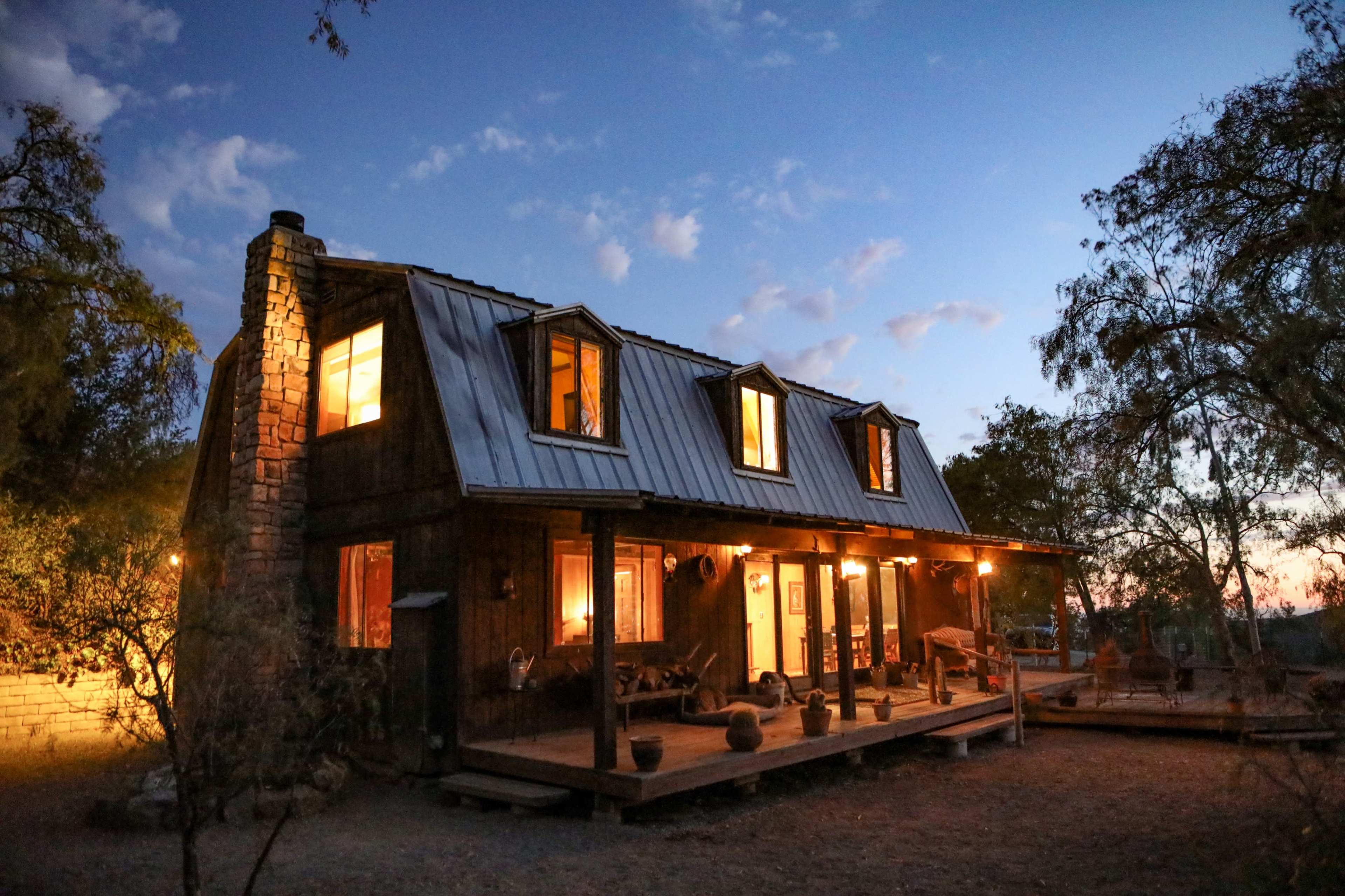 A rustic cabin with a metal roof and illuminated windows stands among trees at dusk.