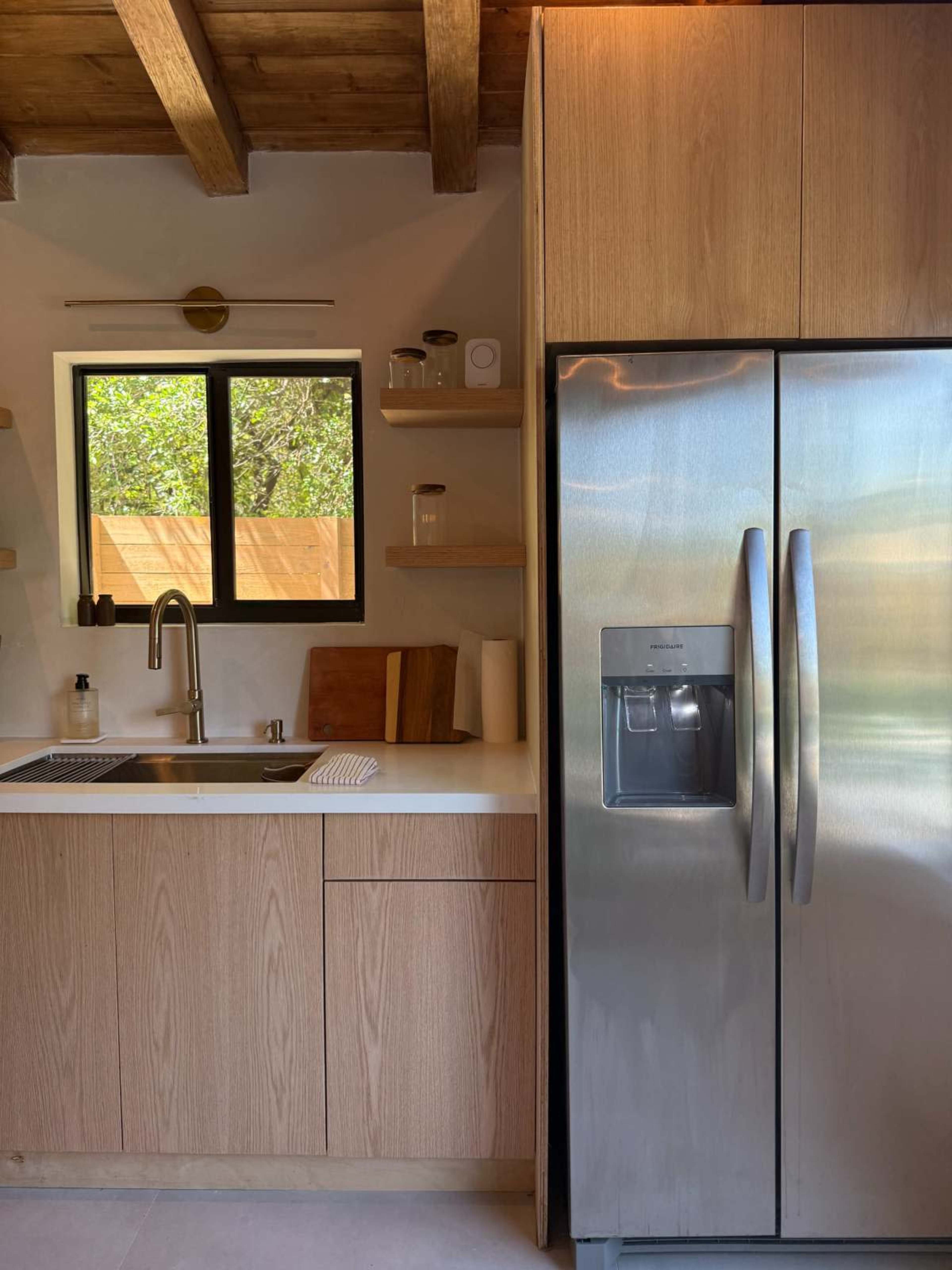 The image shows a modern kitchen with wooden cabinetry, a stainless steel refrigerator, and a sink with a single faucet, complemented by open shelving.