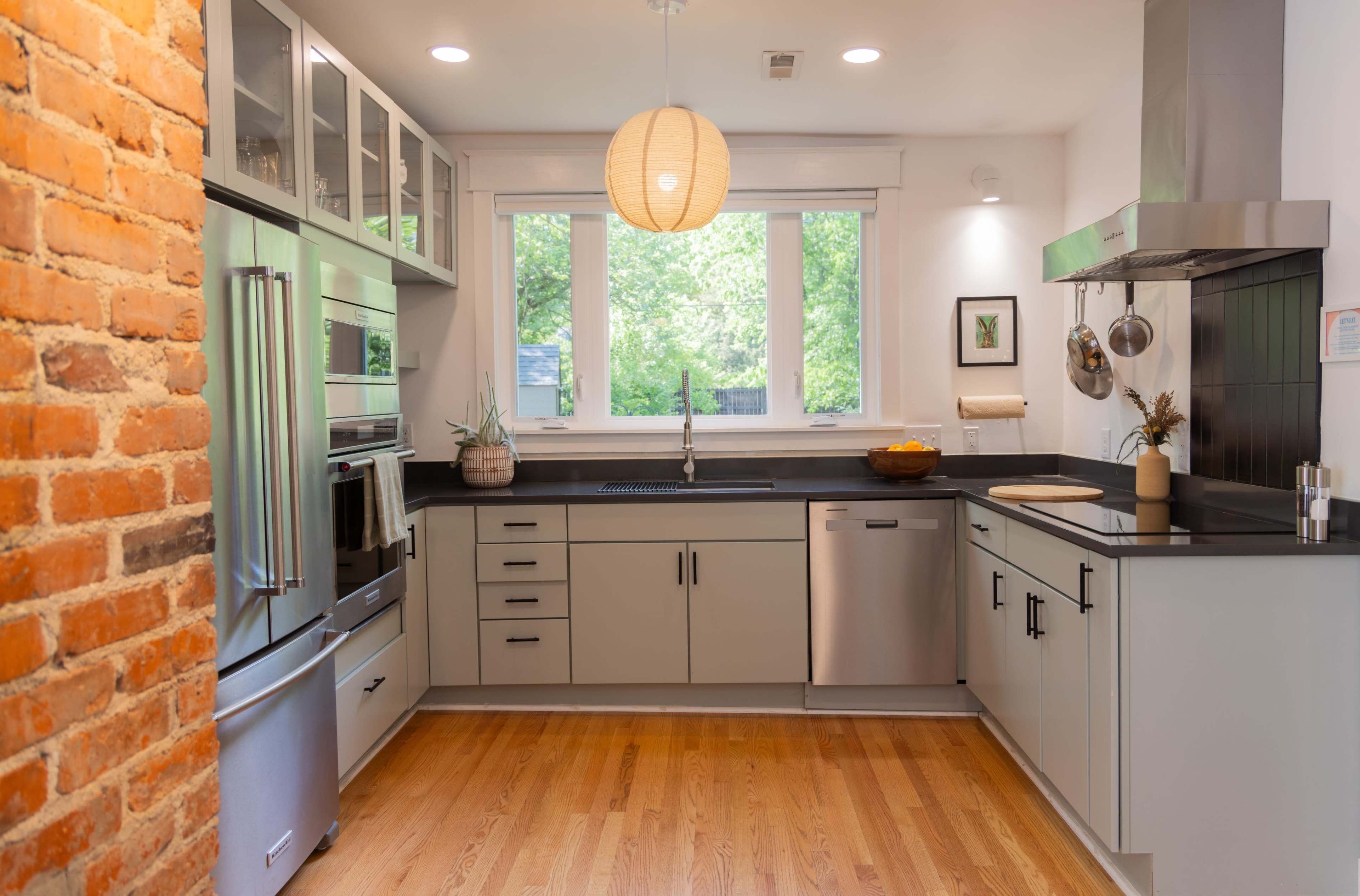 The image shows a modern kitchen with a brick accent wall, stainless steel appliances, gray cabinetry, and wooden flooring.