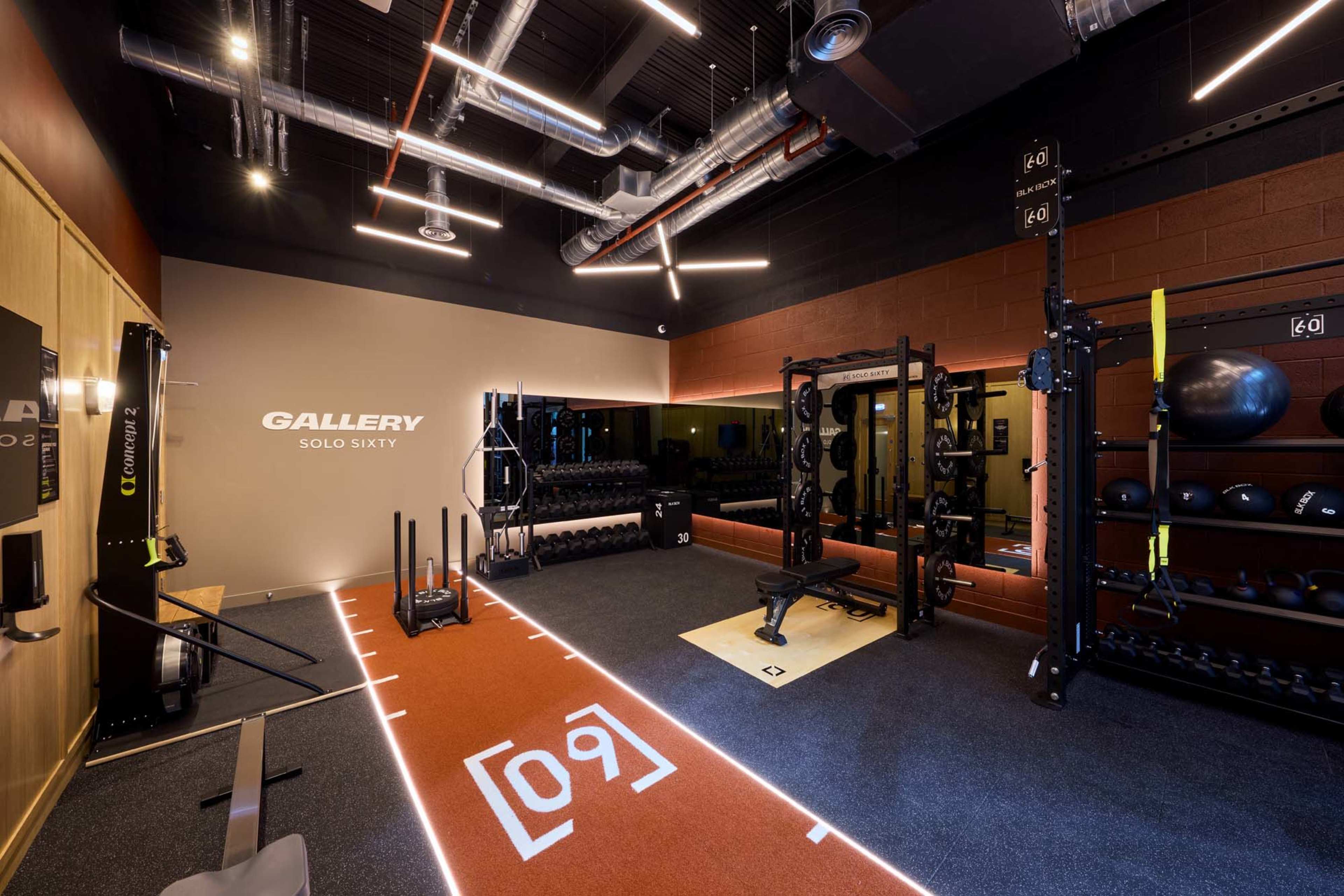 The image shows a modern fitness gym with an area for weightlifting, exercise equipment, and a mirrored wall.