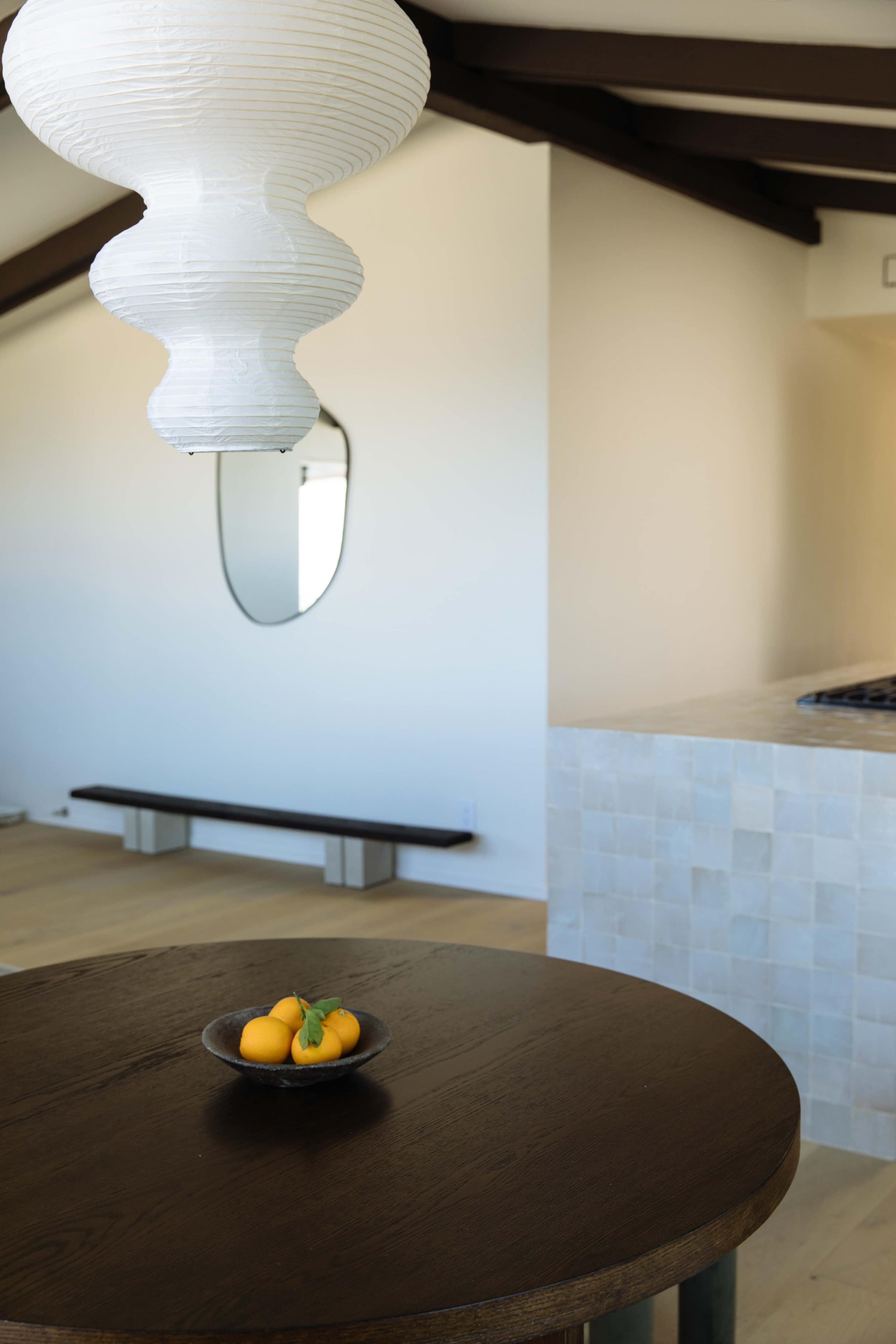 A round wooden table with a bowl of oranges is set under a white pendant light in a minimalistic room with a kitchen counter and a mirror on the wall.