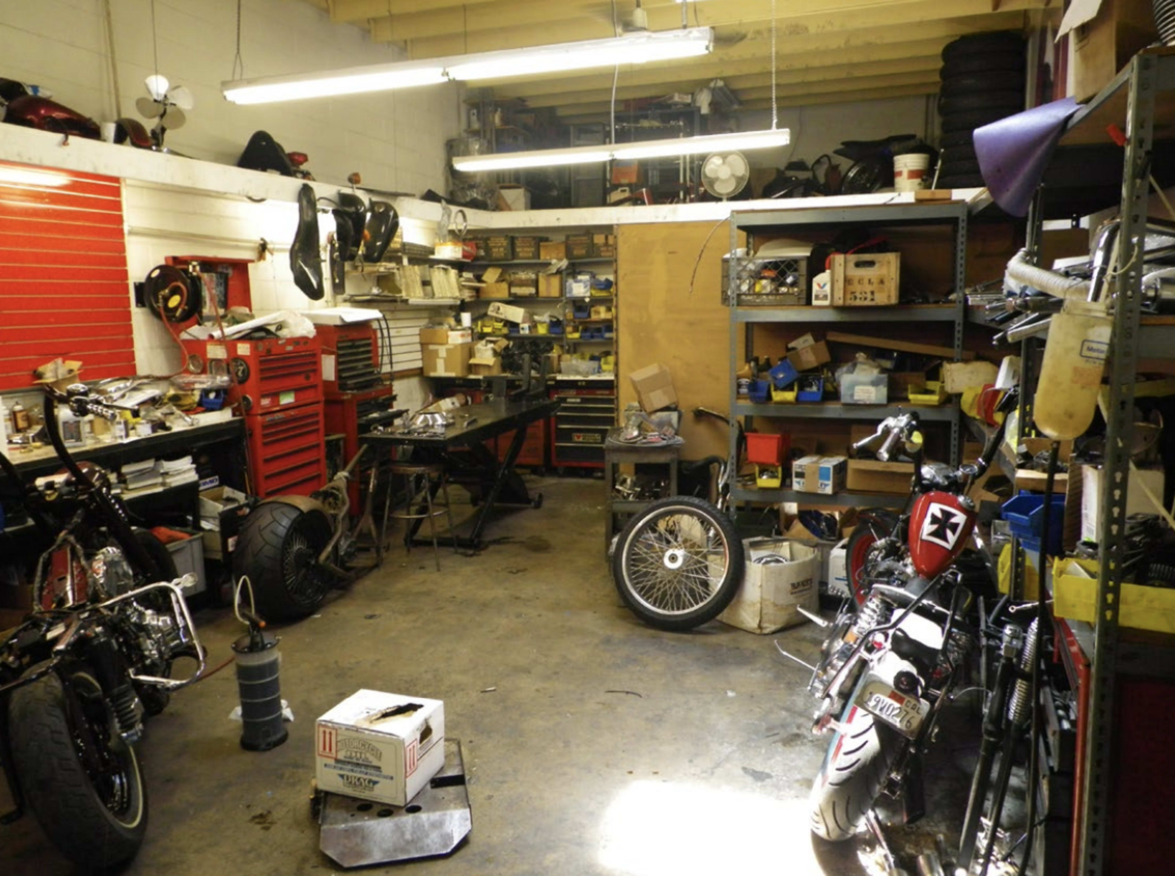 The image shows a cluttered garage workspace with various motorcycle parts, tools, and shelves filled with equipment.