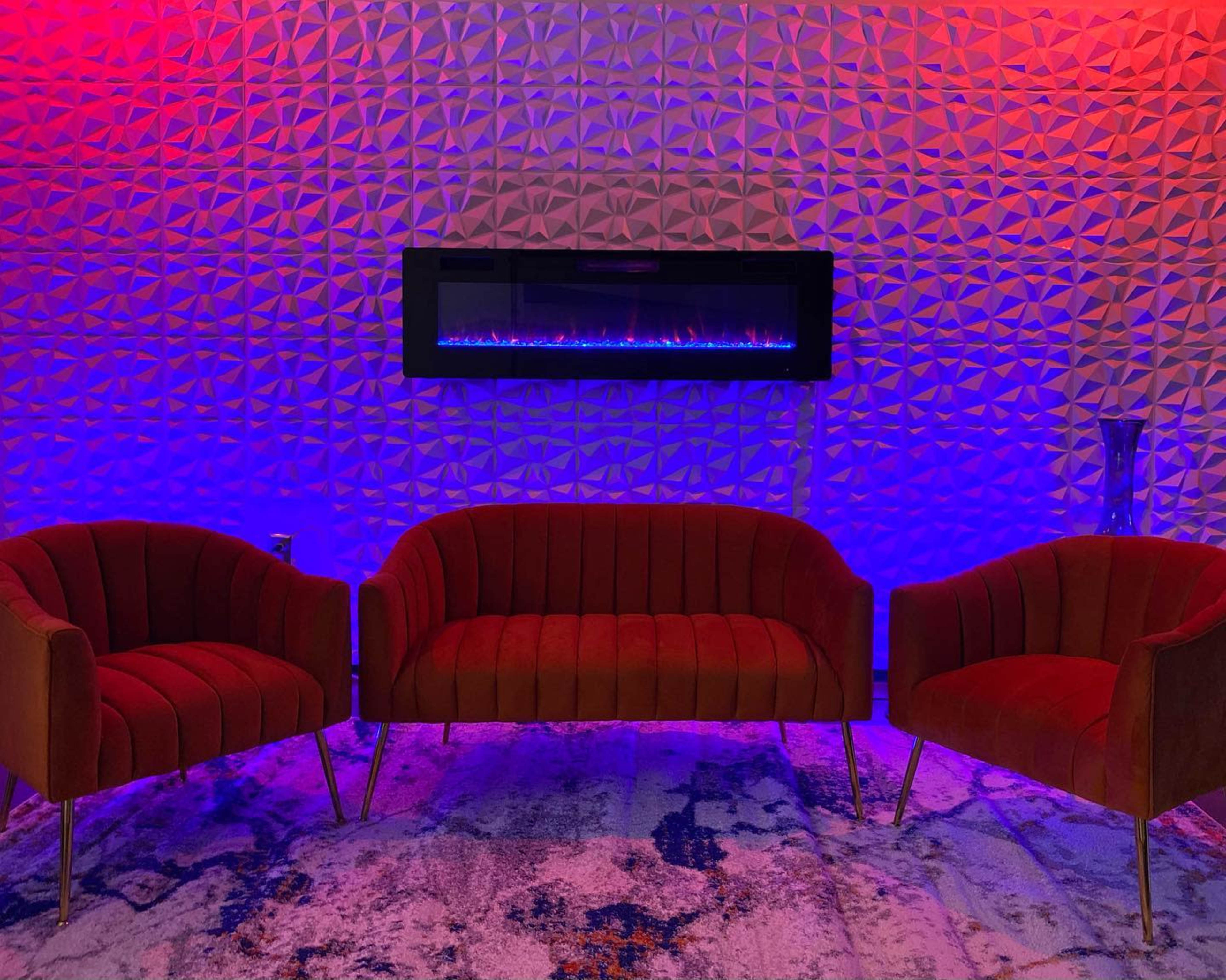 The scene features three red velvet chairs arranged near a modern wall-mounted electric fireplace, illuminated with blue and red lights against a textured wall.