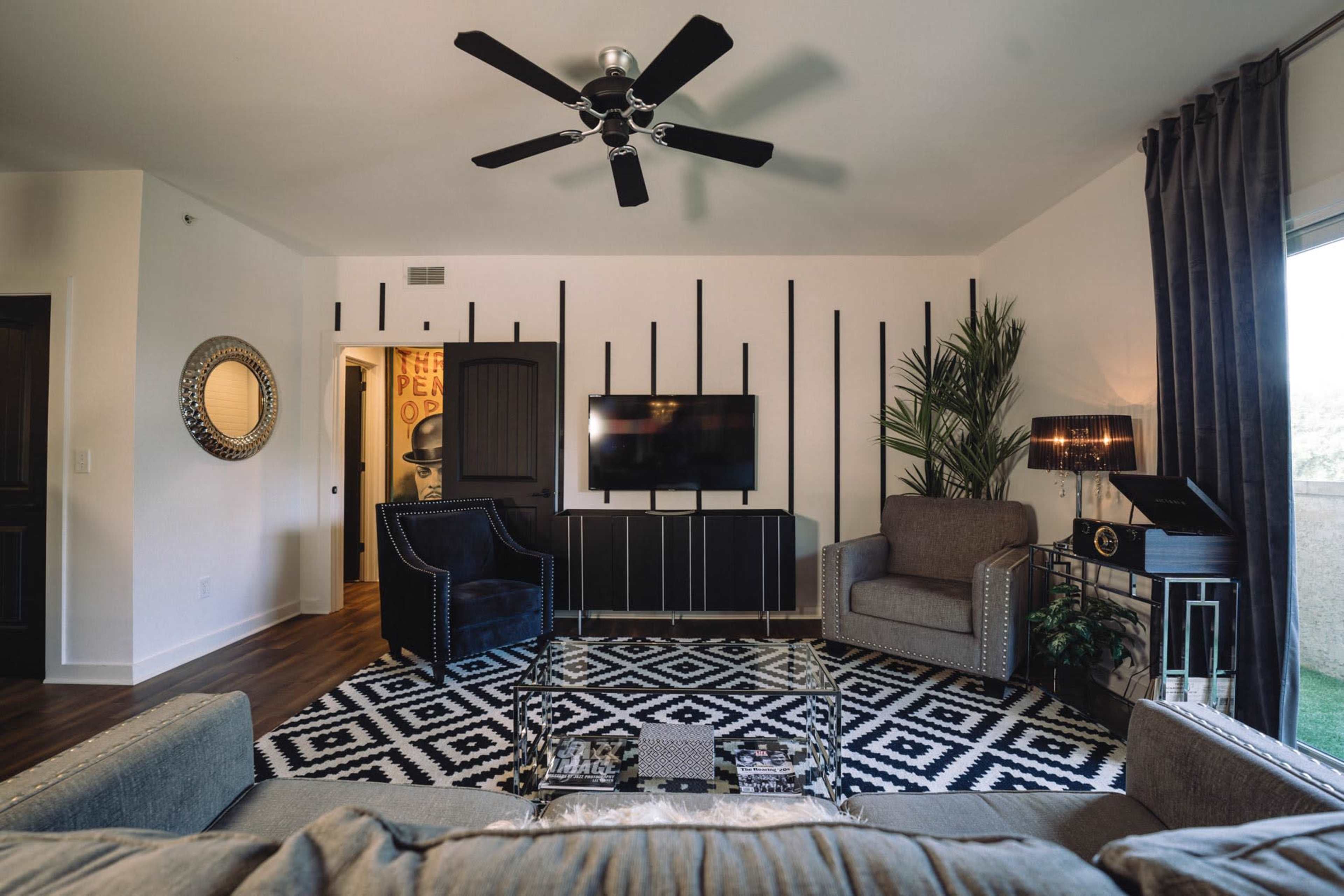 A modern living room features a large television mounted on a striped accent wall, with two armchairs, a coffee table, and a patterned area rug.