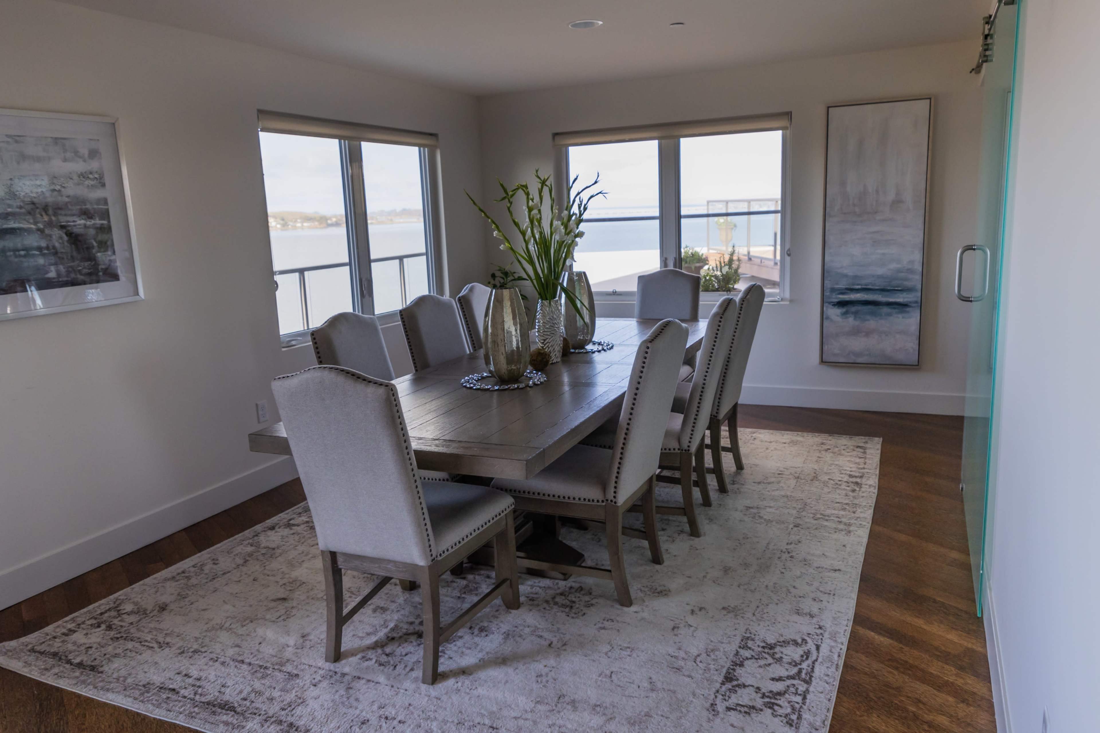 A dining room features a large wooden table surrounded by eight upholstered chairs, with large windows overlooking a body of water.
