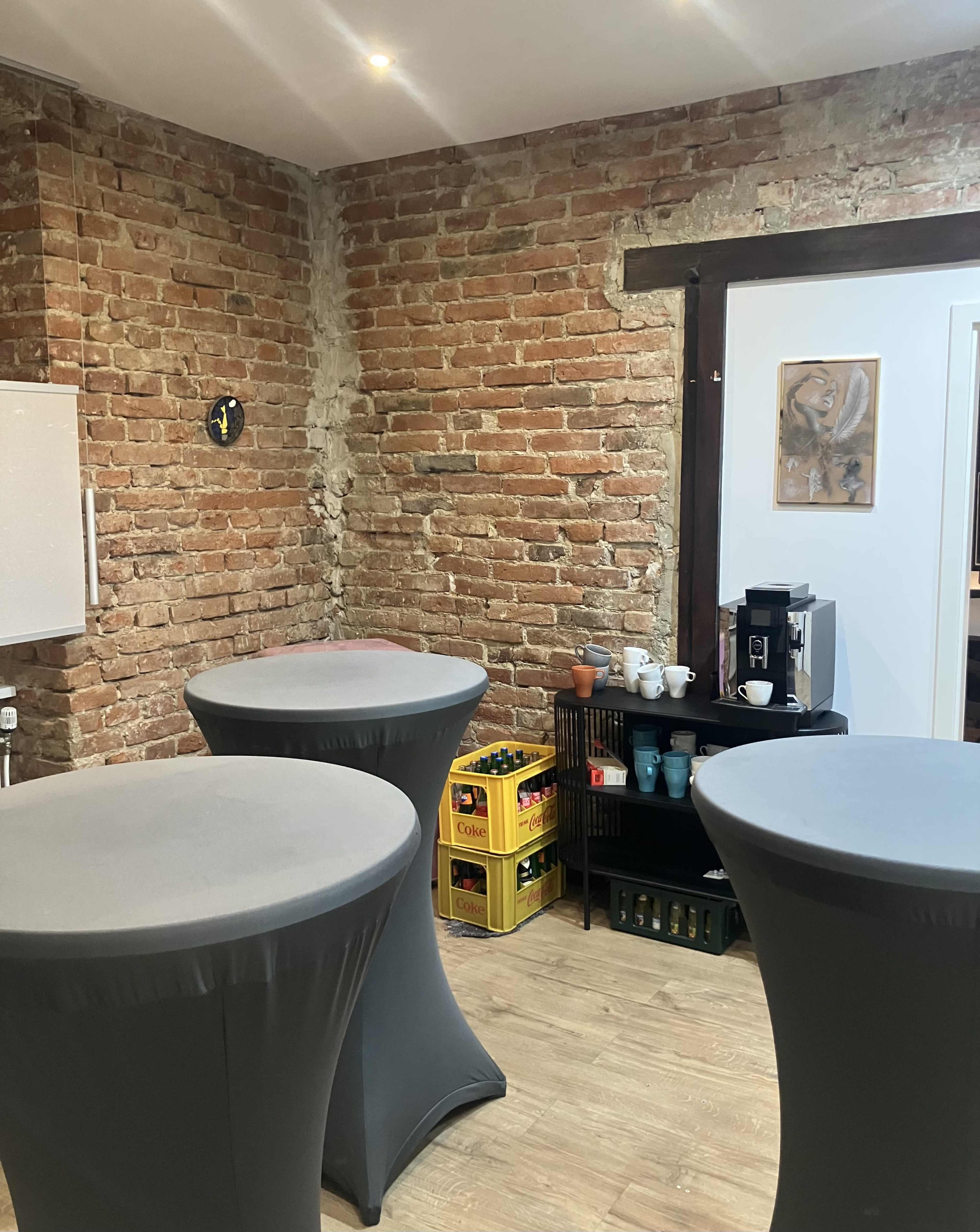 The image shows a room with exposed brick walls, three high tables with gray covers, and a small kitchenette area containing drinks and supplies.