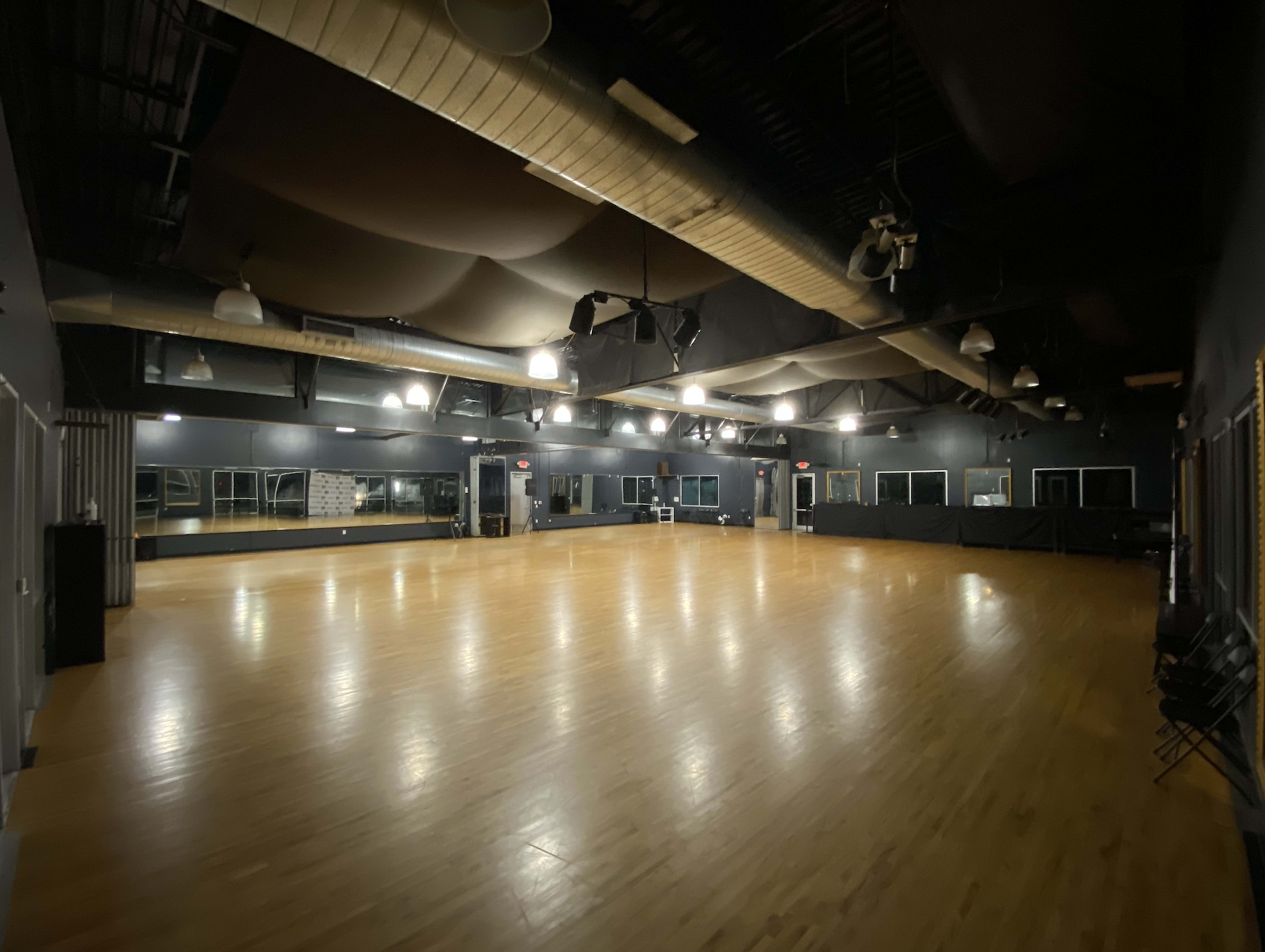 The image shows a spacious dance studio with polished wooden floors, dim lighting, and large mirrors along one wall.