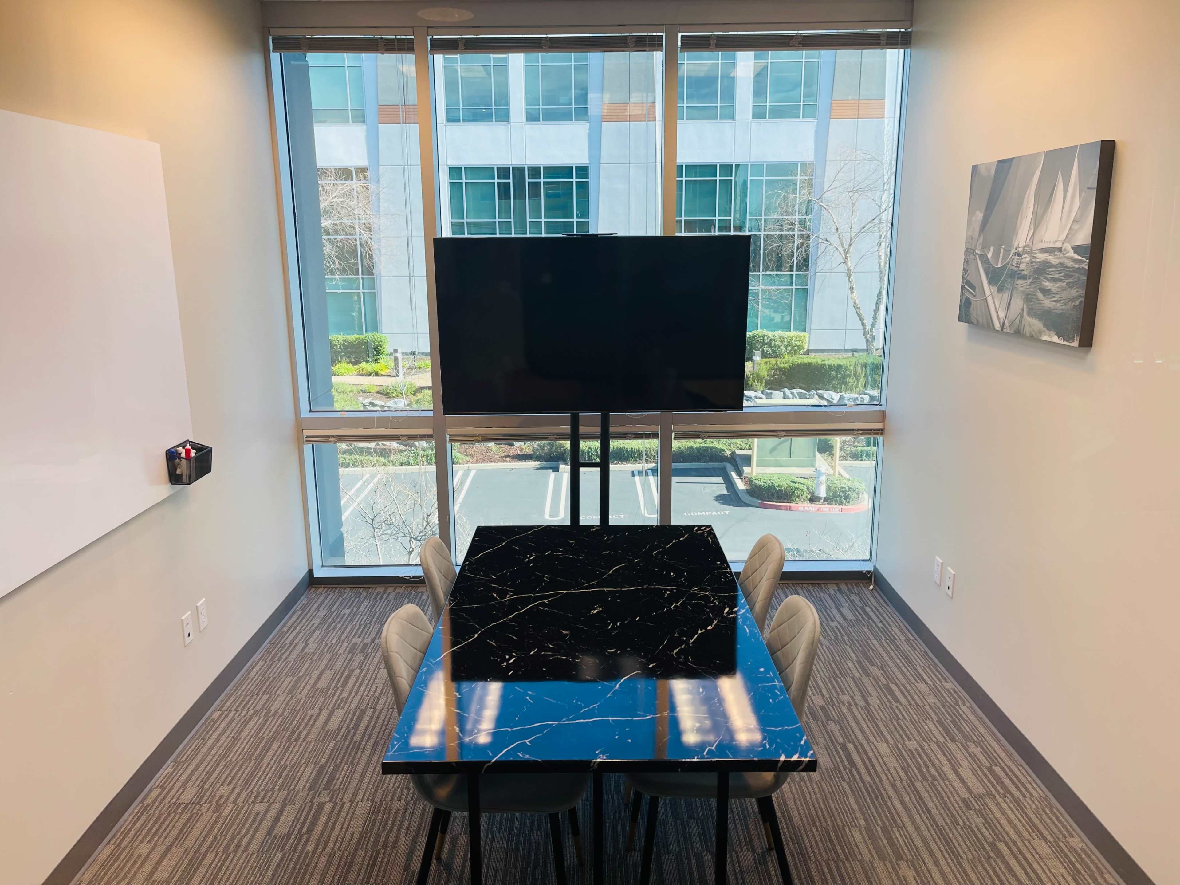 A modern conference room features a black marble table surrounded by light-colored chairs, a large TV on a stand, and large windows providing a view of the outdoors.