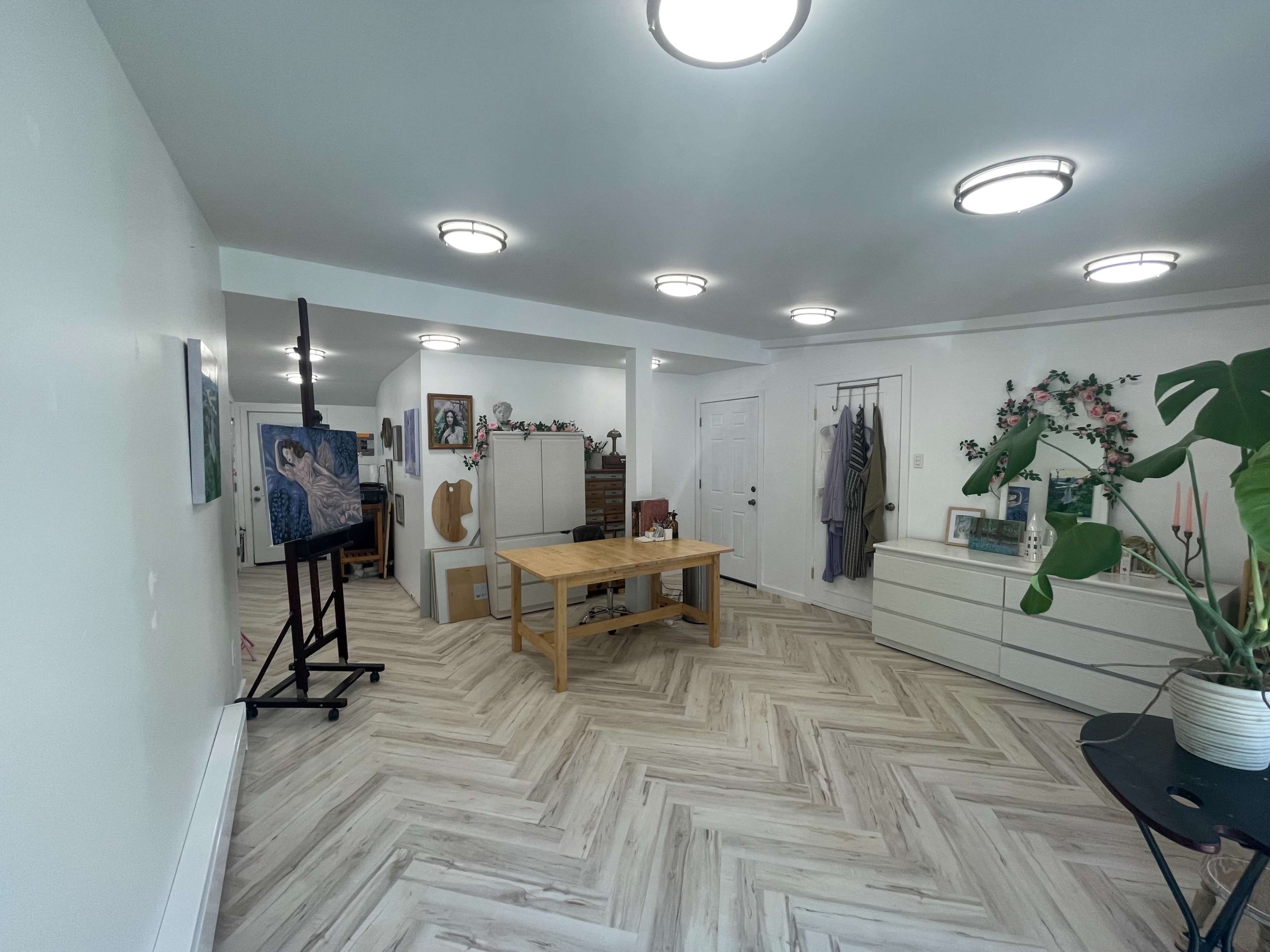 The image shows a spacious, well-lit room featuring a central wooden table, an easel with a painting, several storage units, and decorative plants.