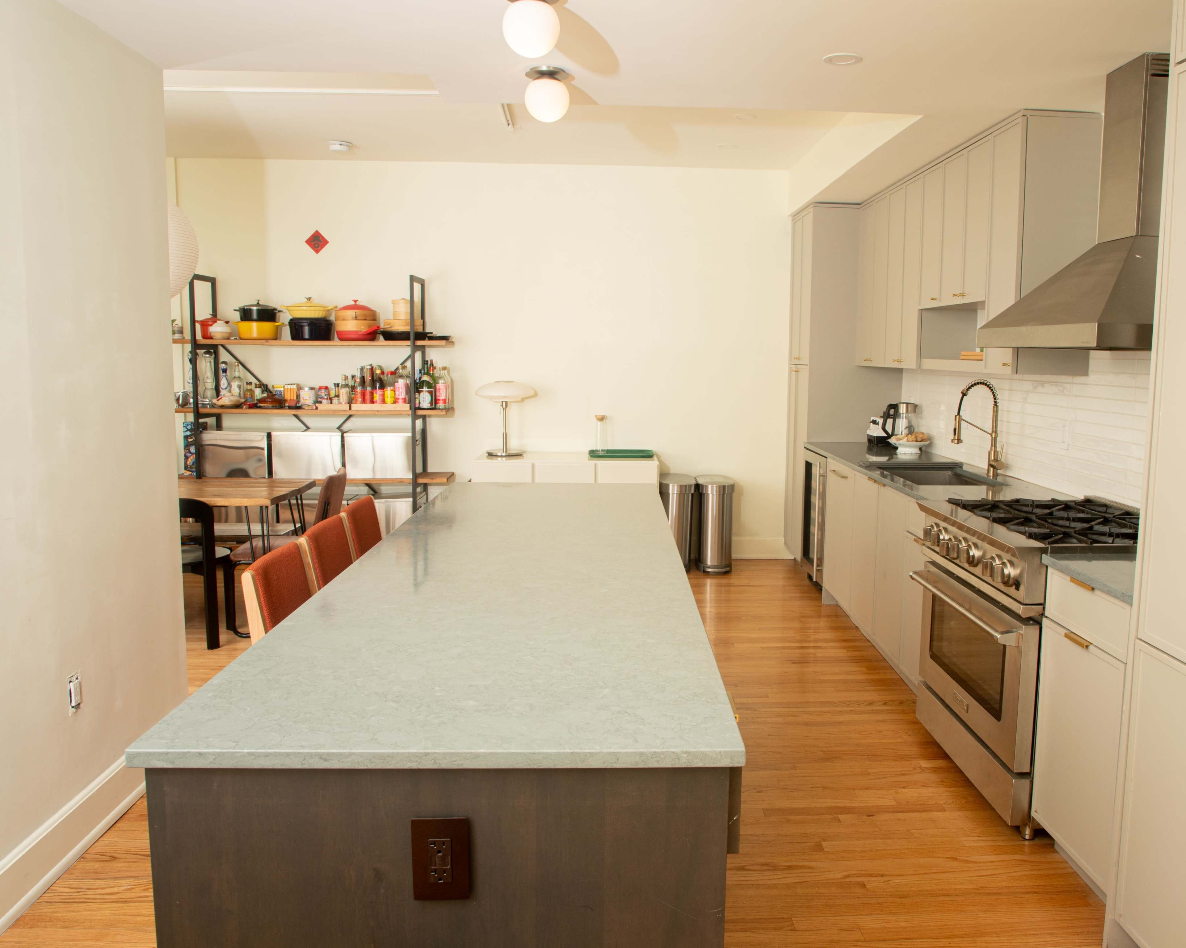 The image shows a modern kitchen with a long central island, stainless steel appliances, and a dining area with wooden chairs.