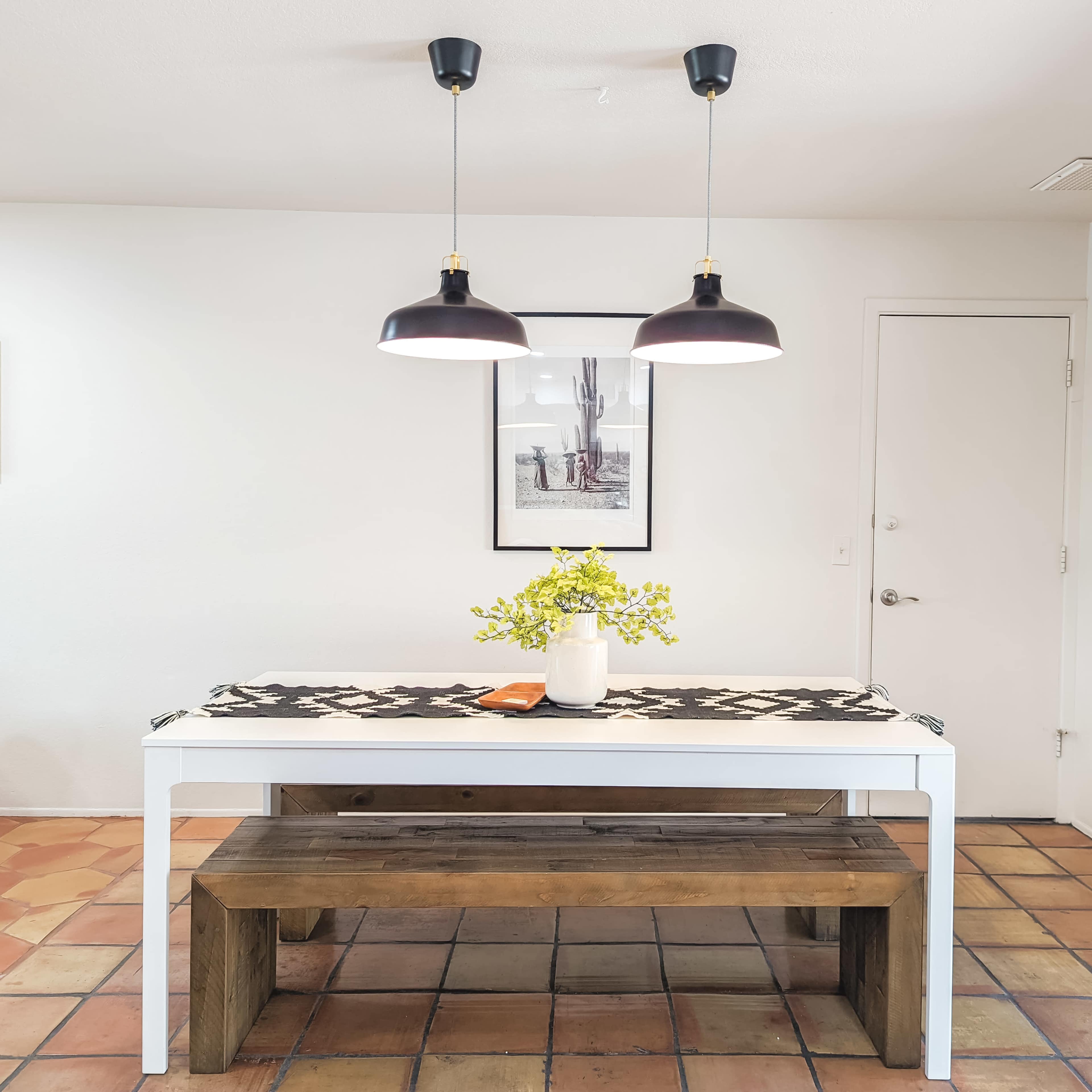 The image shows a minimalist dining area with a table, a wooden bench, a floral centerpiece, and pendant lights hanging above.