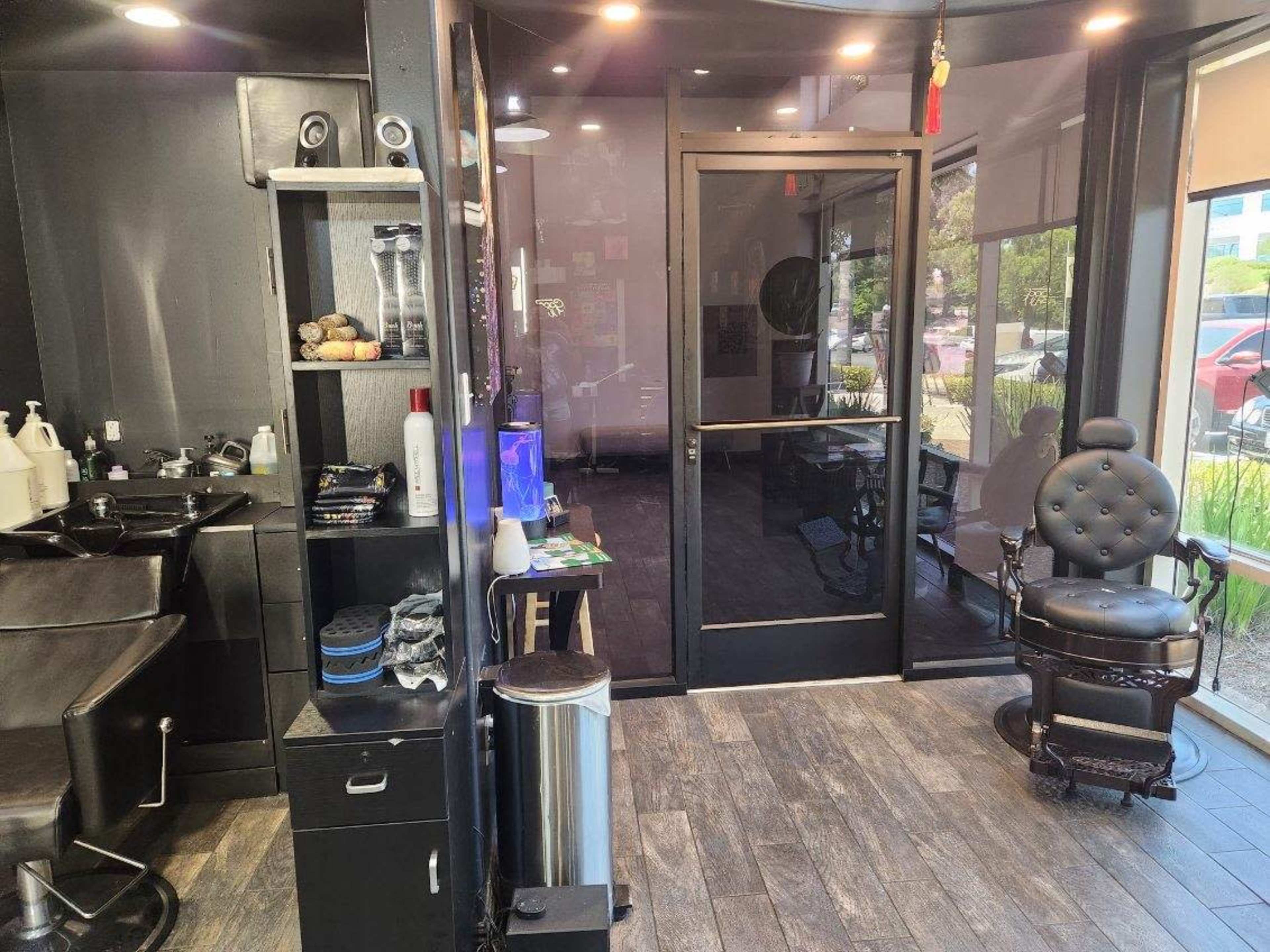 The image shows a sleek barbershop interior featuring two black barber chairs, a retail shelving unit with grooming products, and a glass entrance door.
