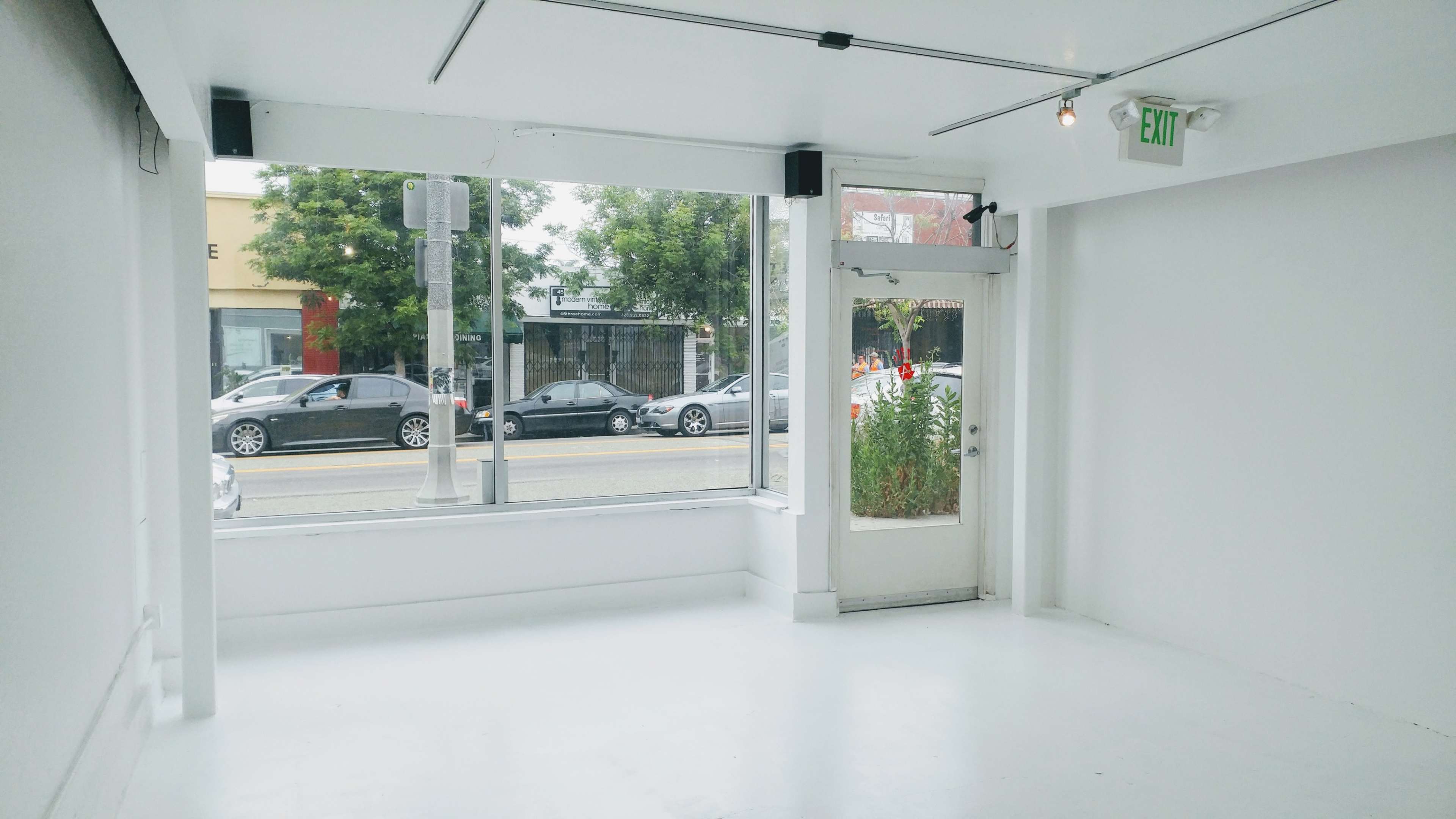 The image shows a bright, empty interior space with large windows overlooking a street and a door on the right.