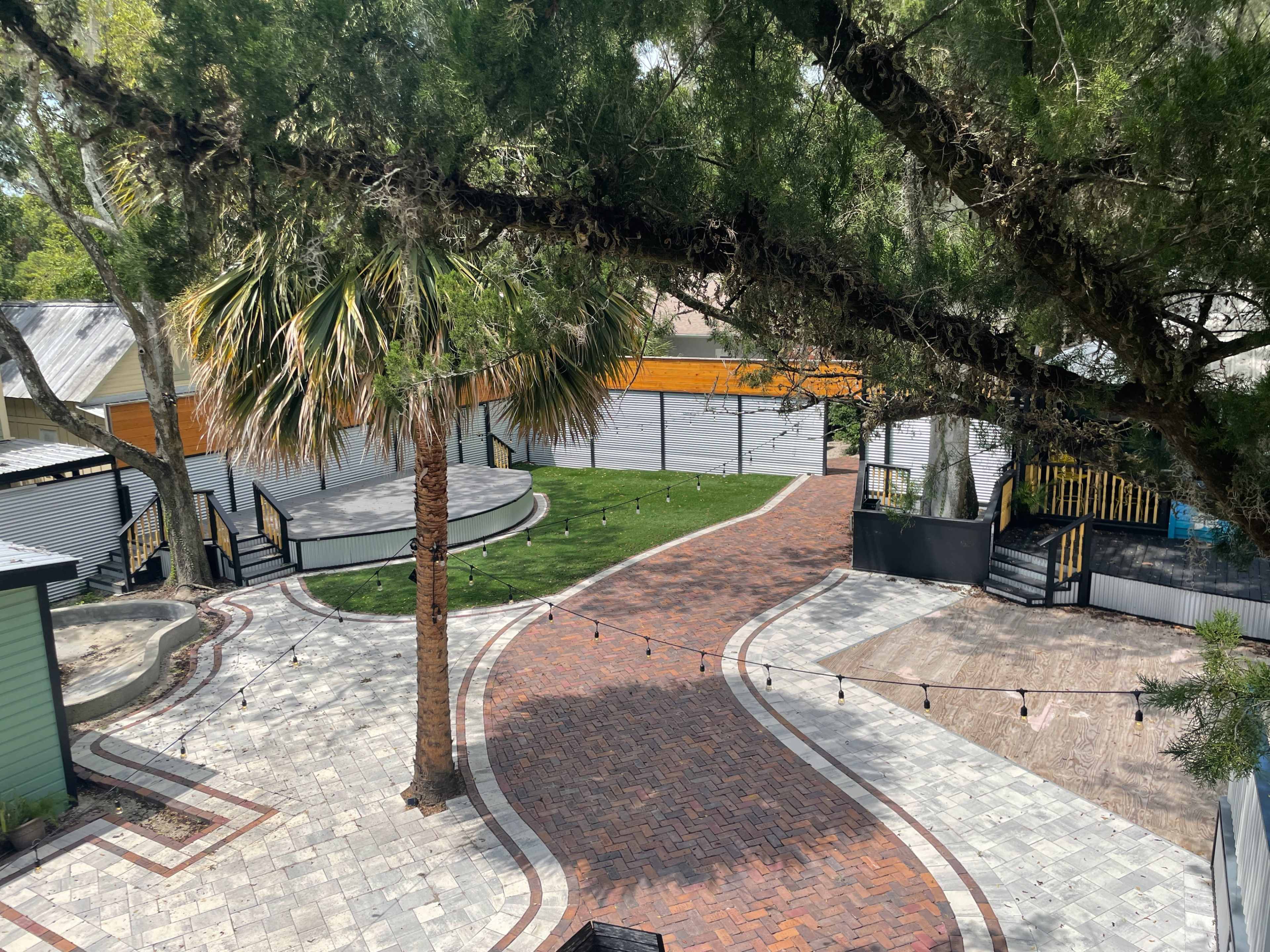 The image shows a landscaped outdoor area featuring winding brick pathways, palm trees, and various structures including patios and shaded seating areas.