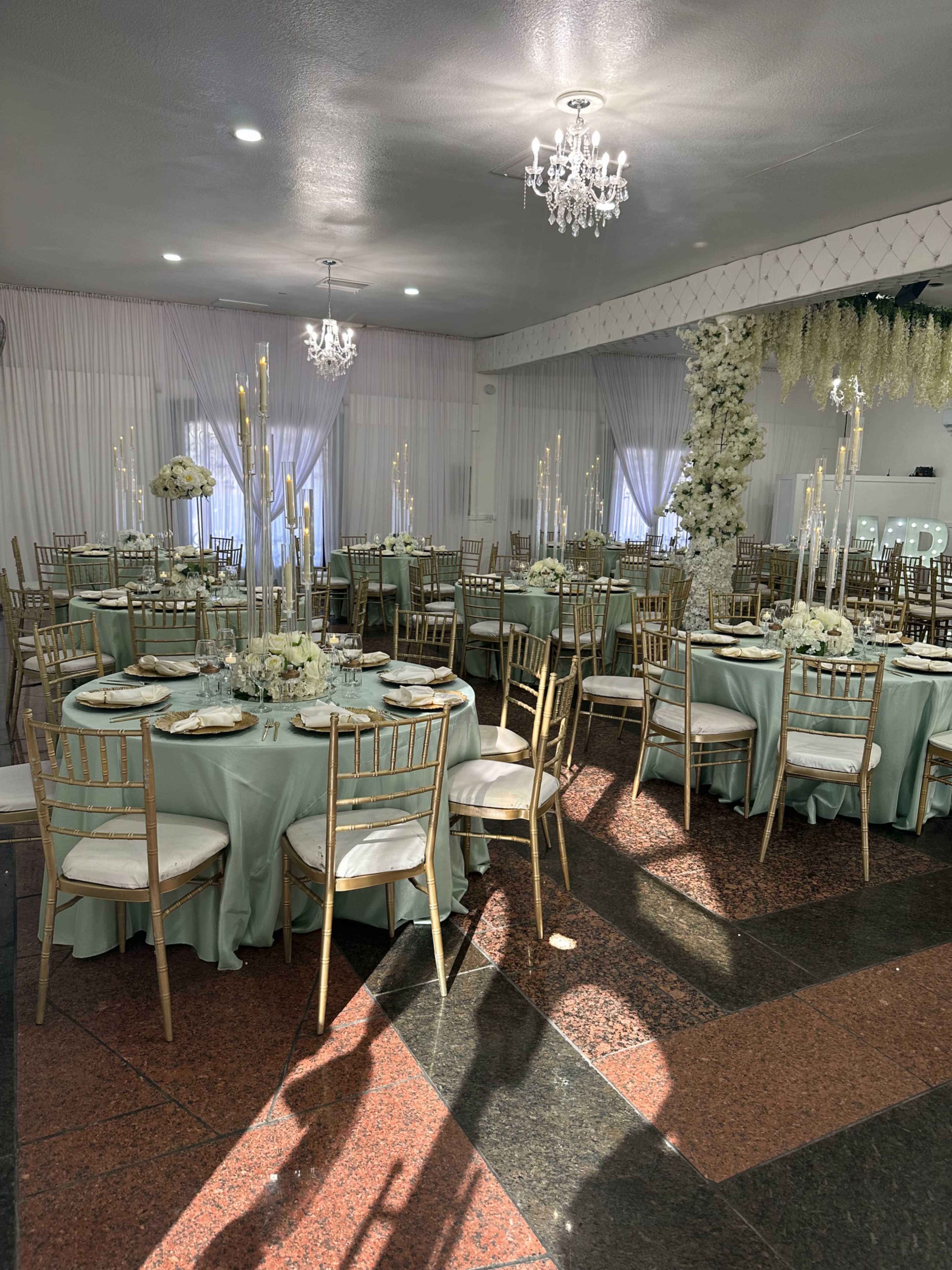 The image shows a banquet hall set up with round tables dressed in light blue tablecloths and gold chairs, featuring tall candle holders and floral centerpieces, illuminated by chandeliers.