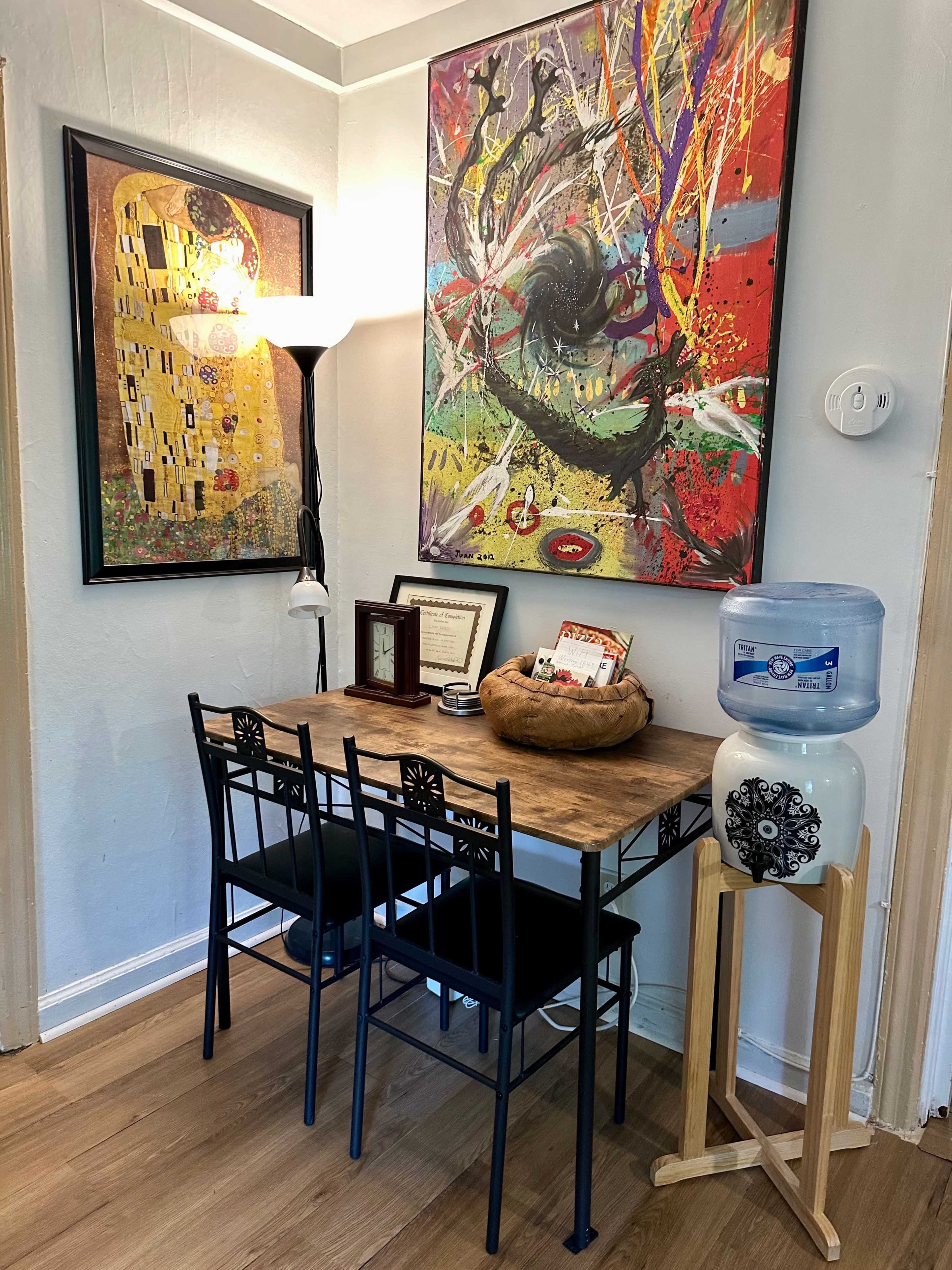 A small dining area features a wooden table with four black chairs, a decorative bowl on the table, and two colorful paintings on the wall above, alongside a water cooler in the corner.