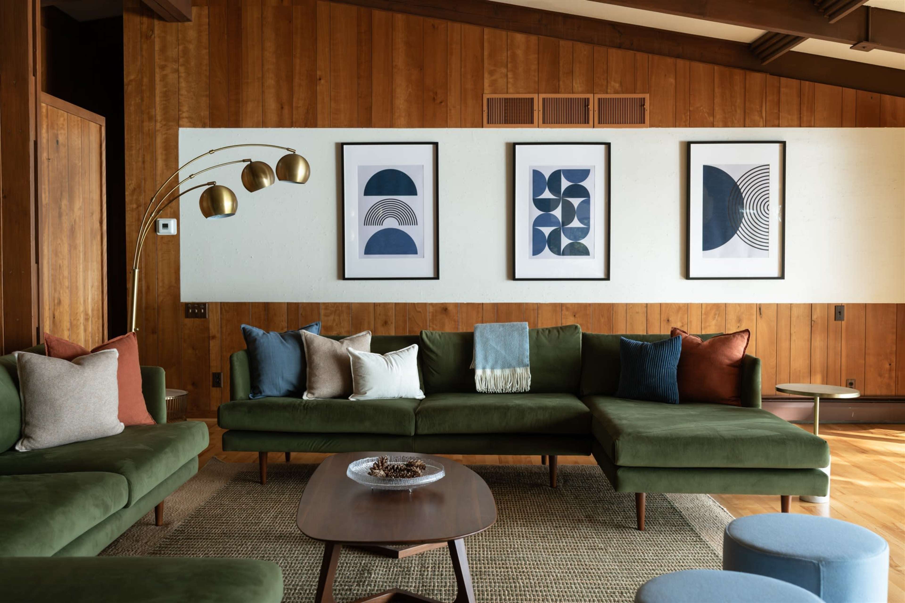 The image shows a modern living room with a green sectional sofa, a wooden accent wall, framed geometric artwork, and various decorative pillows.