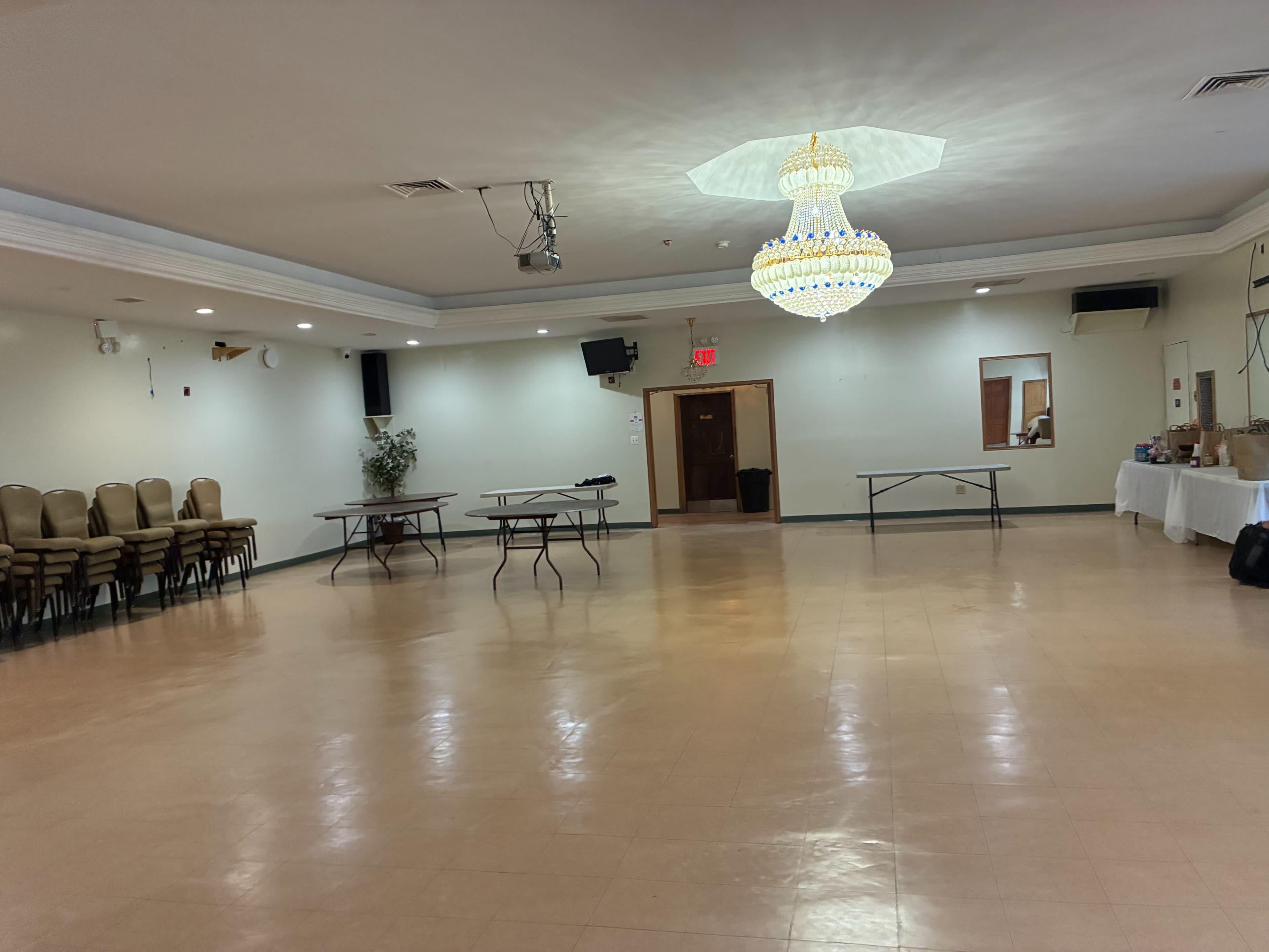 The image shows a spacious, empty event hall with several tables and chairs lined against the wall, and a chandelier hanging from the ceiling.