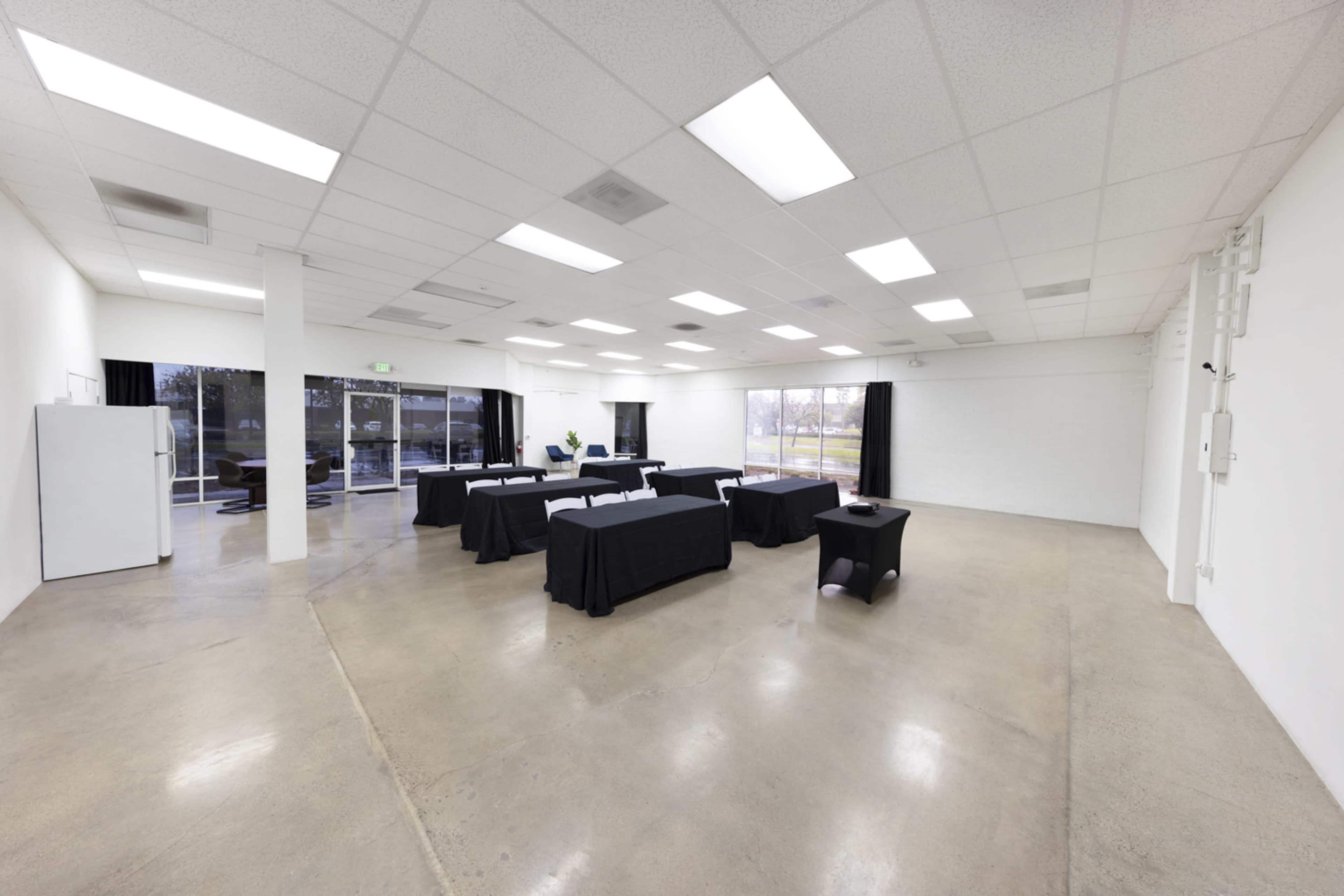 A spacious room with concrete flooring and fluorescent lighting contains several tables covered in black tablecloths and large windows offering natural light.
