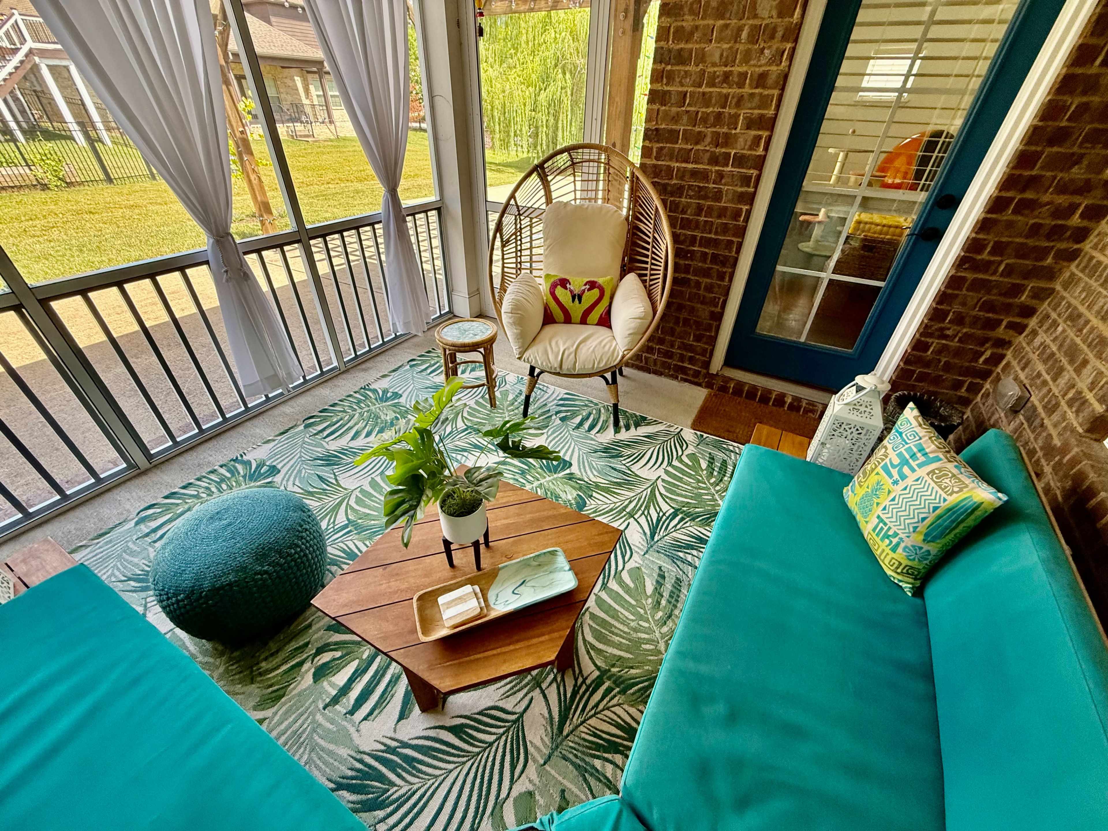 A cozy screened-in porch features a hanging chair, teal sofas, a wooden coffee table, and a vibrant rug with tropical patterns.