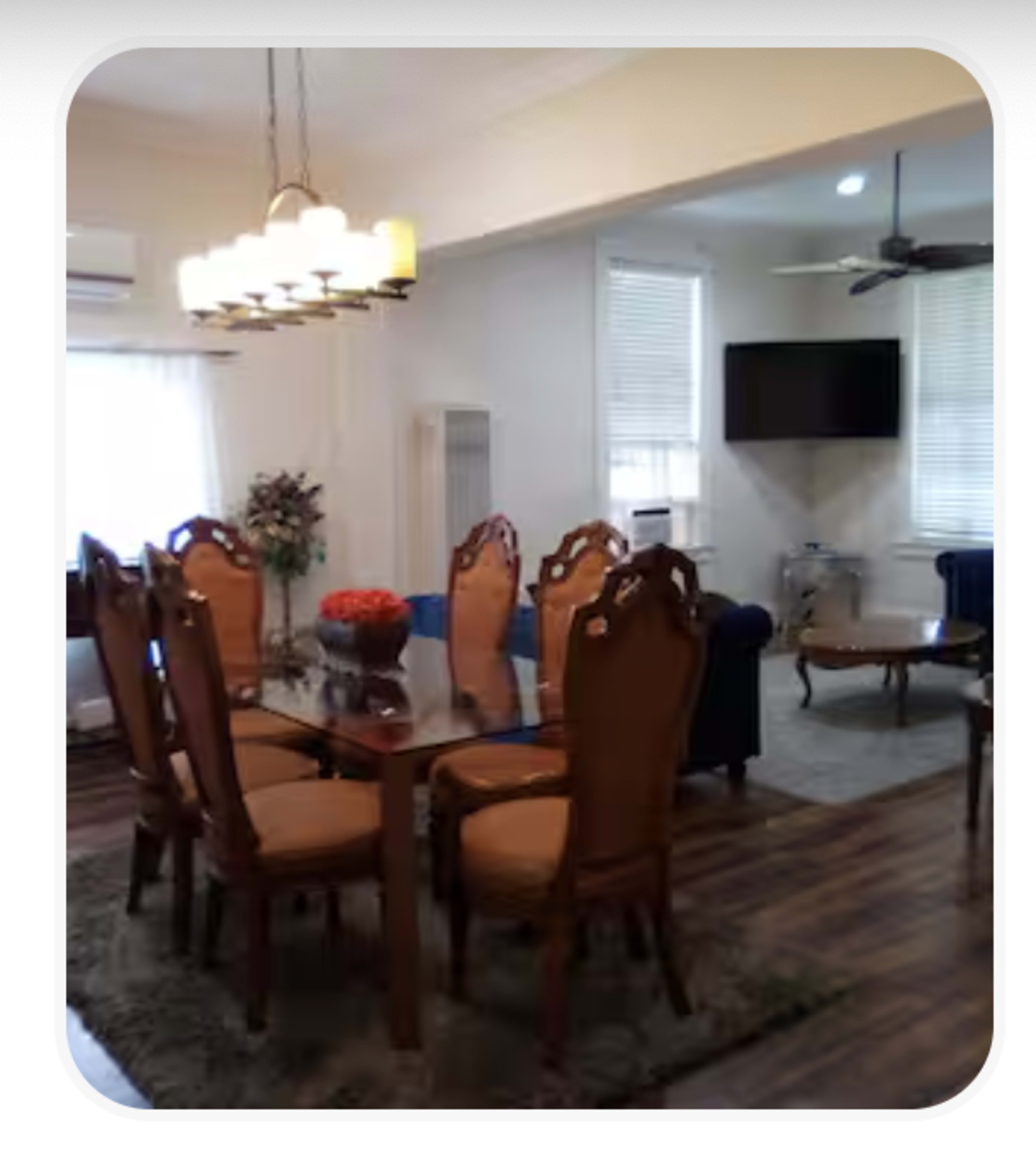 The image shows a dining area with a wooden table and eight chairs, accompanied by a living area featuring a blue couch and a wall-mounted television.