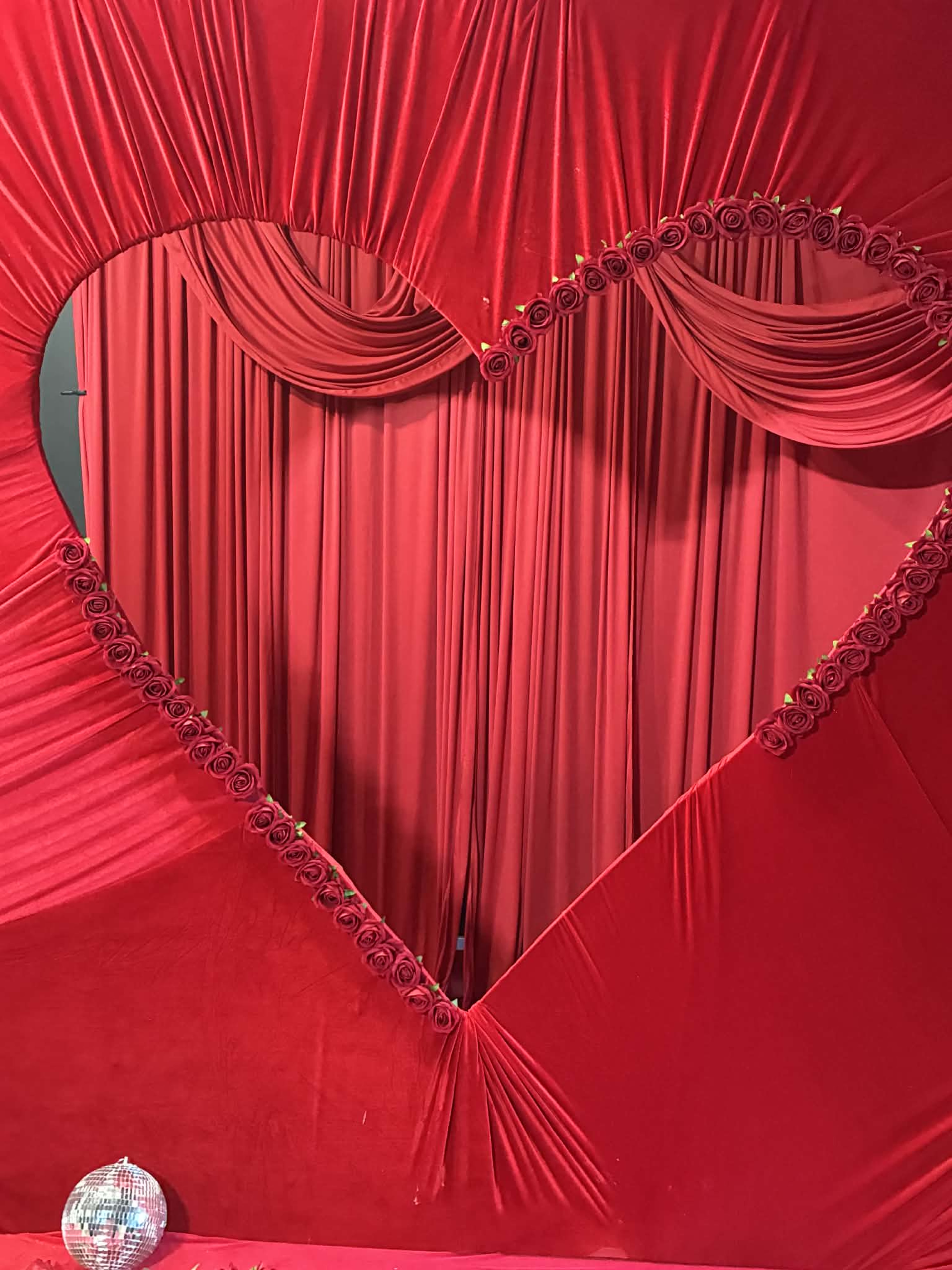 A large red heart-shaped backdrop is decorated with flowing fabric and roses, with a disco ball positioned at the bottom.