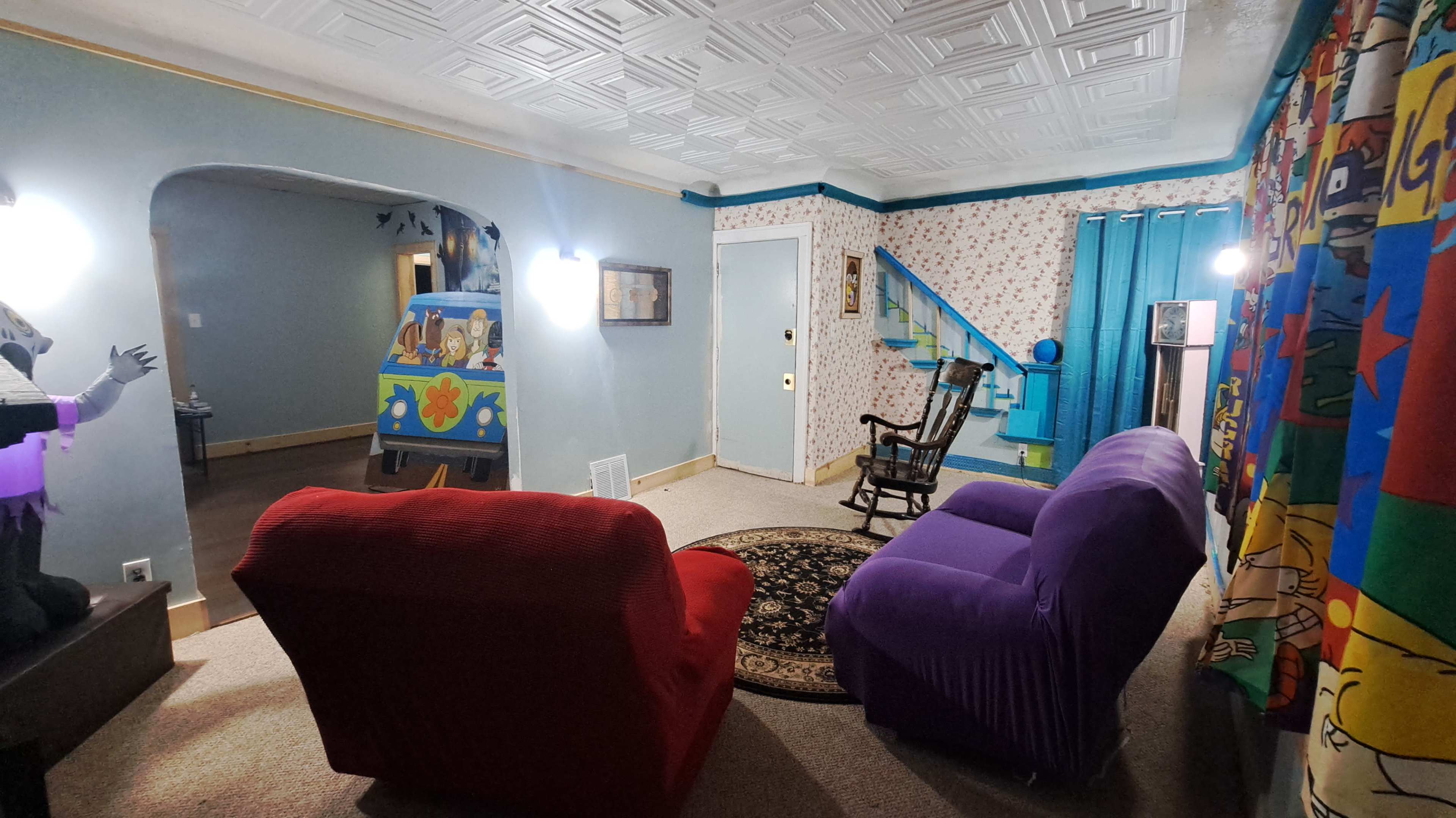 The living room features two colorful armchairs facing a patterned area rug, with blue curtains and floral wallpaper.