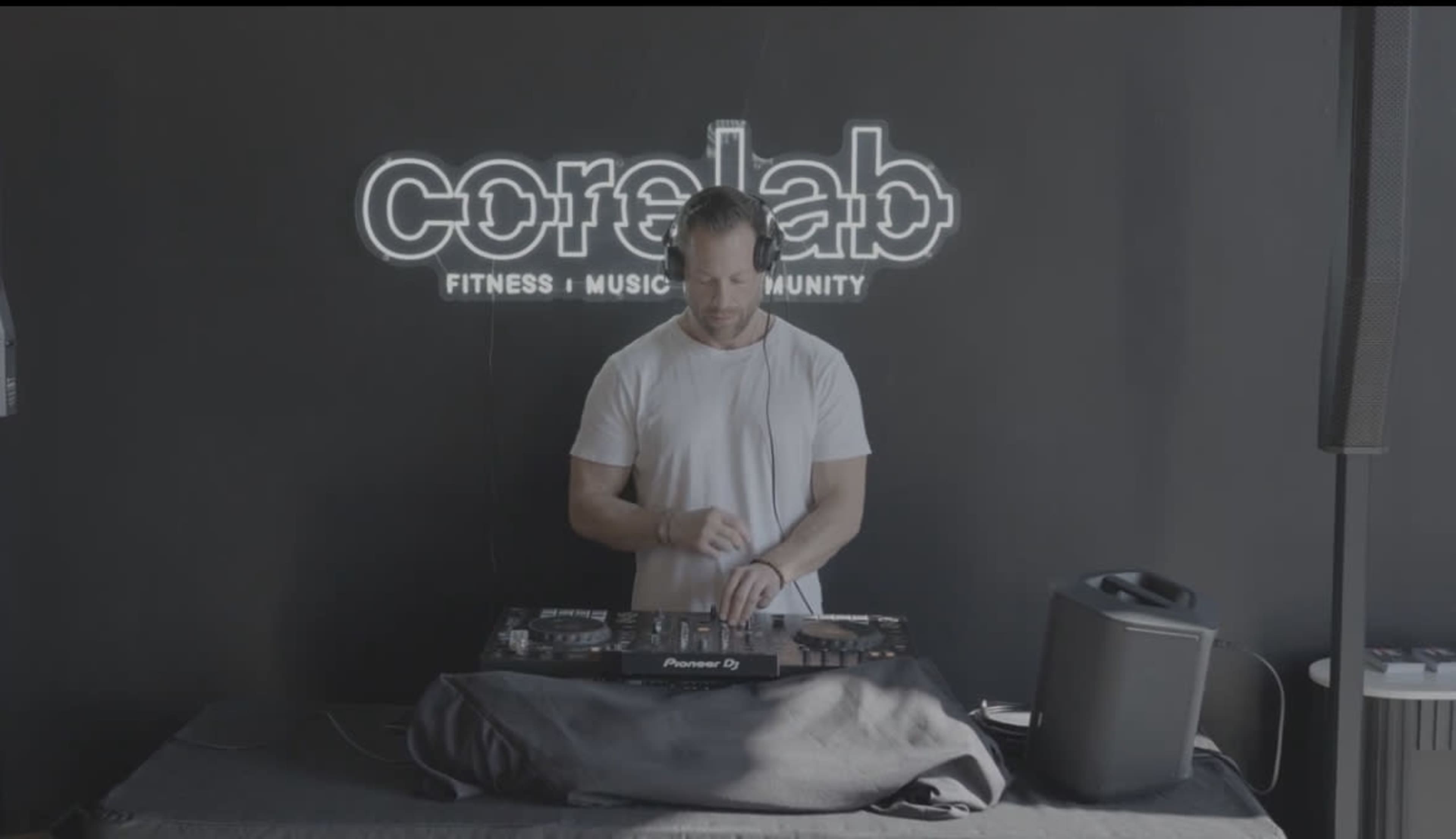 A DJ stands behind a music mixer in front of a black wall featuring the logo of CoreLab, which focuses on fitness, music, and community.