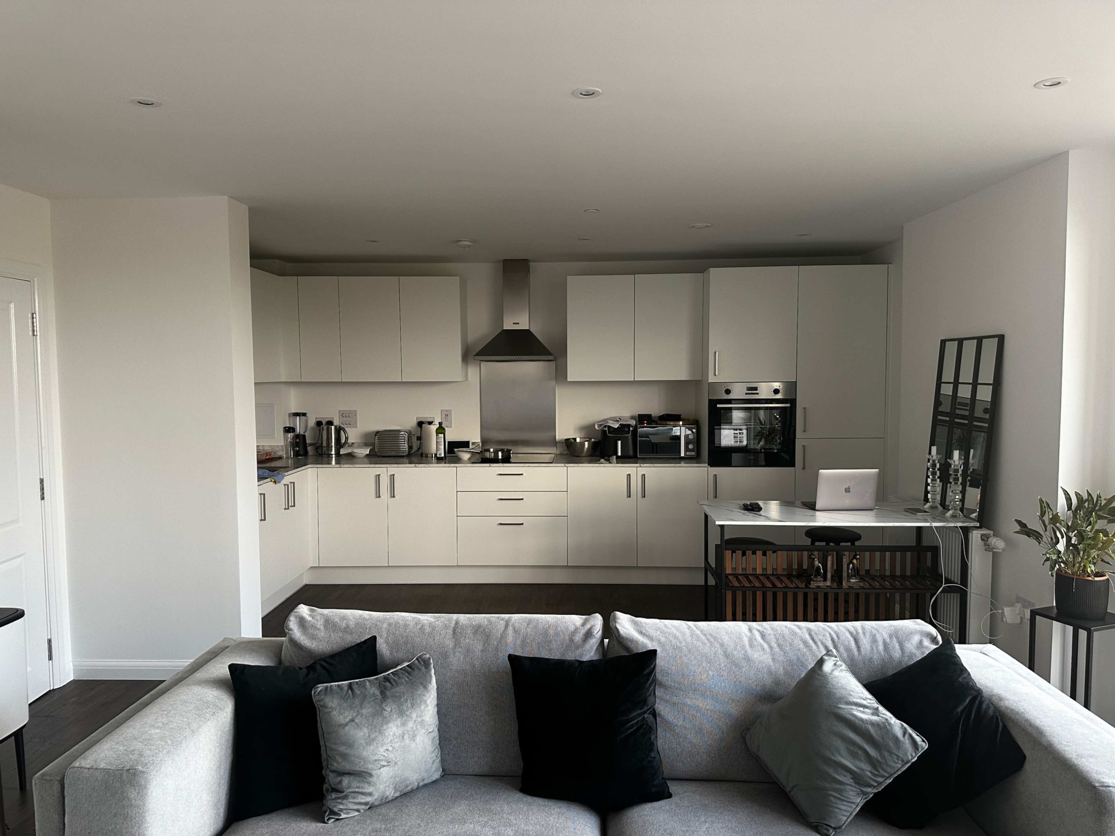 A modern kitchen featuring white cabinets, stainless steel appliances, and a small dining table with a laptop, viewed from a cozy living area with a gray sofa.