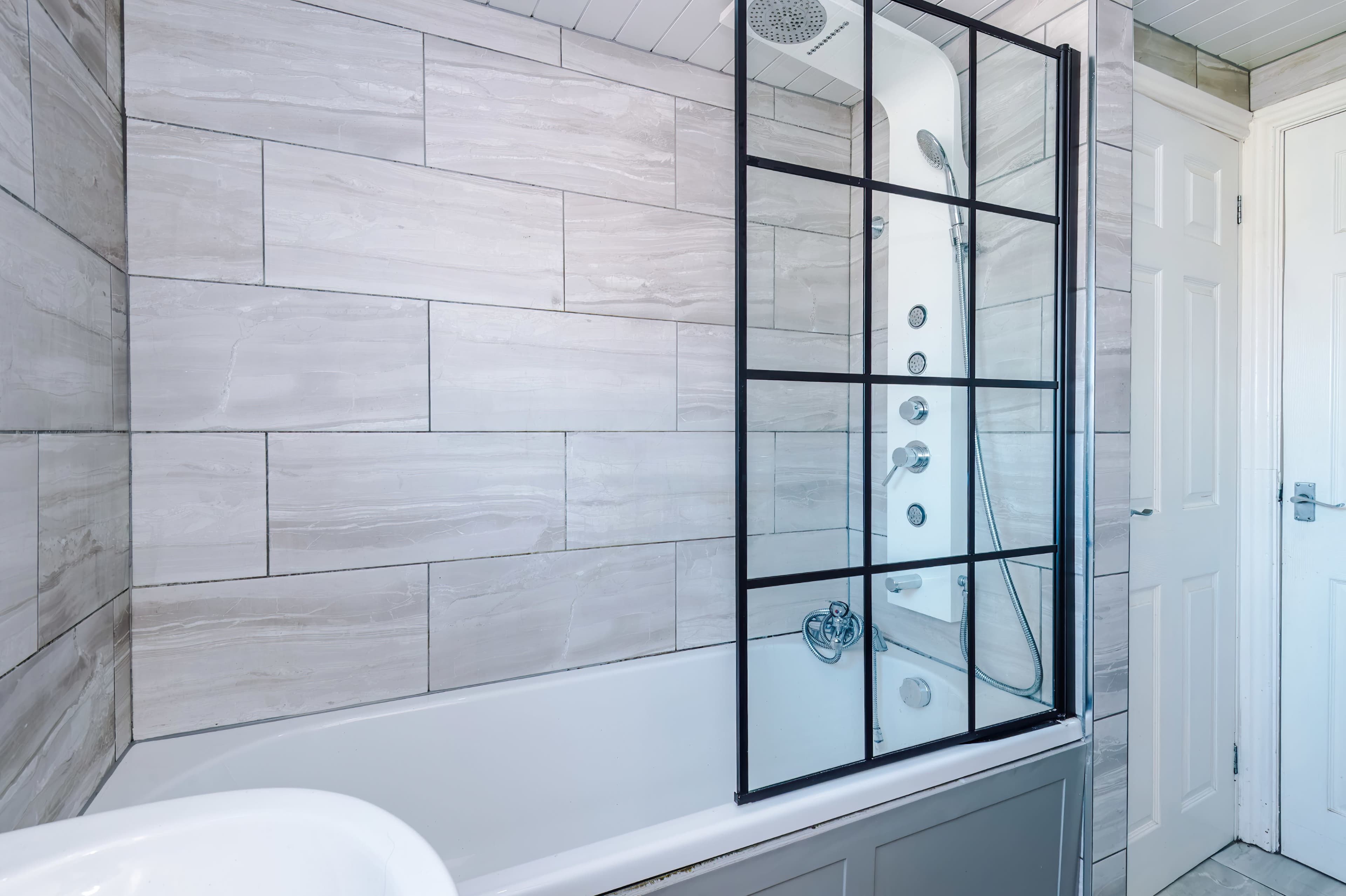 The image shows a modern bathroom featuring a bathtub with a glass shower screen and tiled walls.